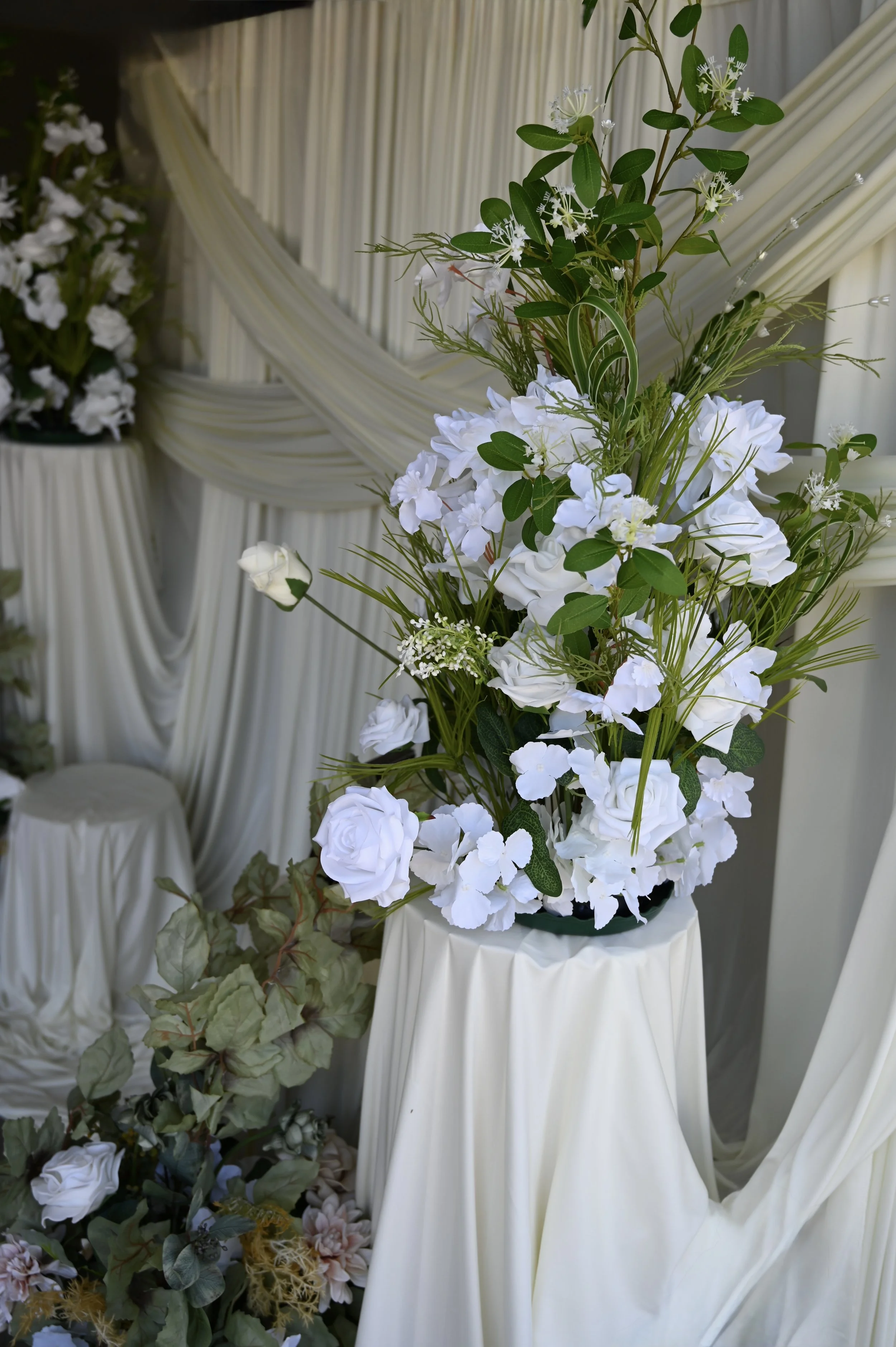 A floral arrangement with white flowers and green leaves, set against a backdrop of white draped fabric.