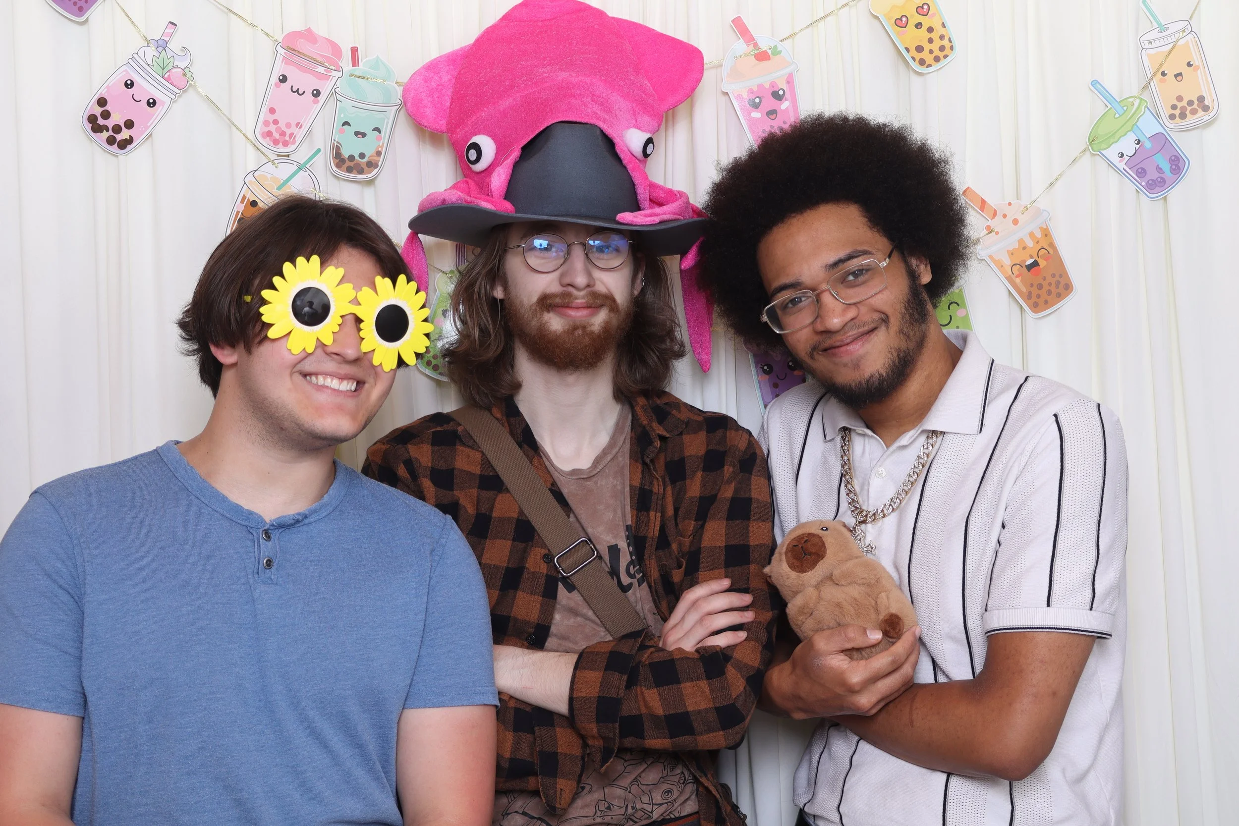 Three men at a fun party, with colorful bubble tea themed decorations and a pink octopus plush hat, wearing playful accessories like sunflower sunglasses, and holding a teddy bear.