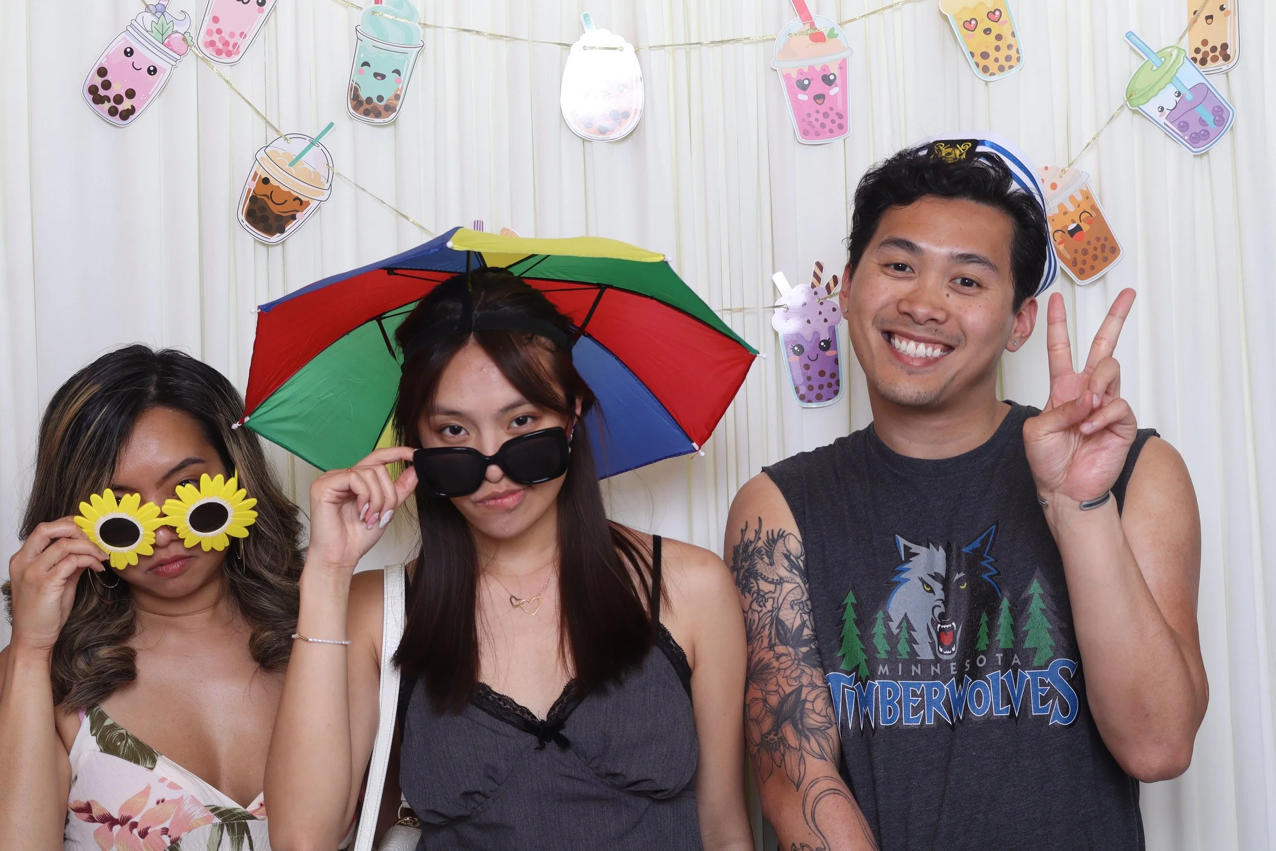 Three people posing in front of a bubble tea-themed backdrop: a woman with sunflower sunglasses, a woman holding an umbrella over her head, and a man flashing a peace sign.