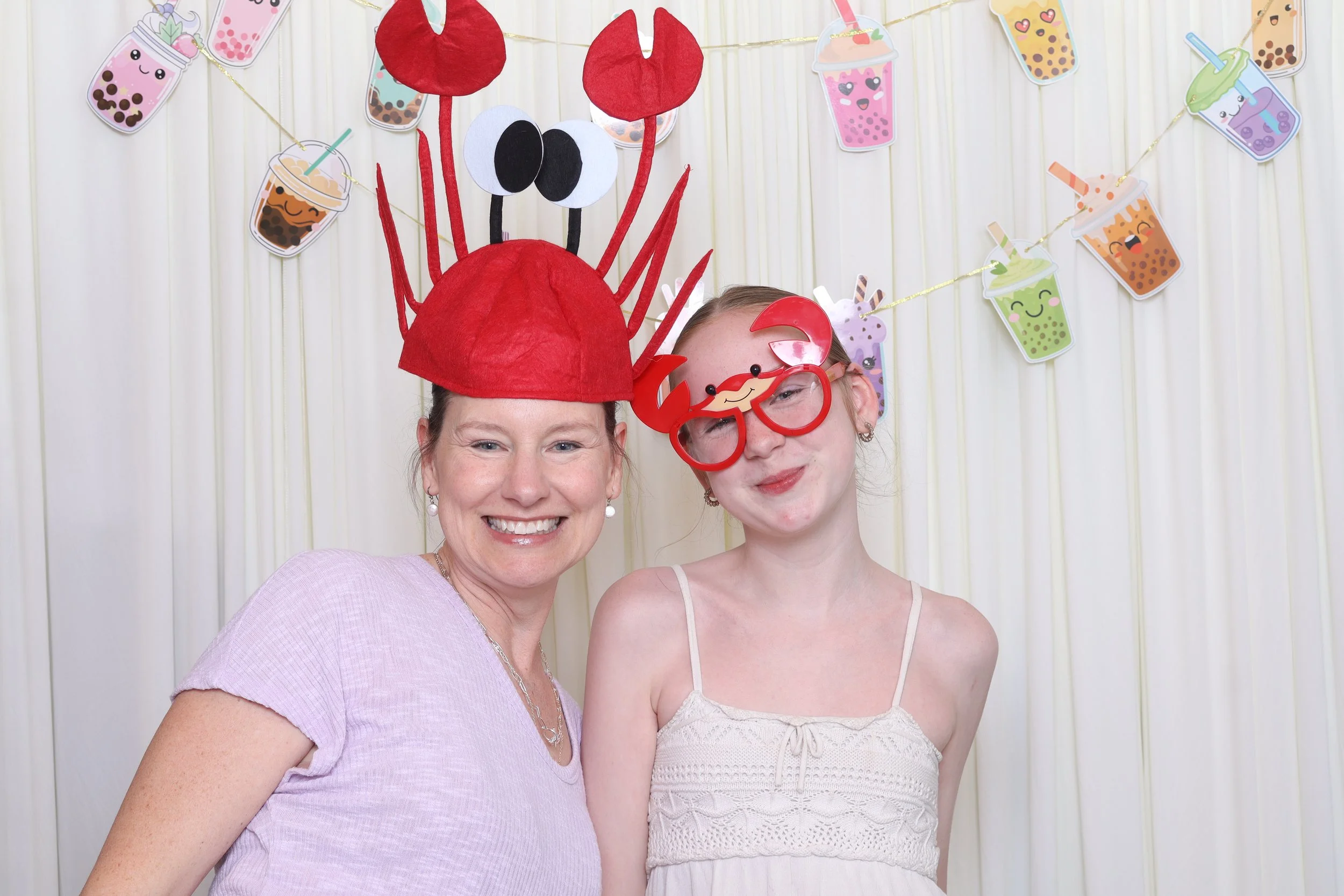 A woman and a girl celebrating a birthday with fun accessories. The woman is wearing a red paper lobster hat and the girl is wearing novelty crab glasses. They are smiling in front of a cream-colored curtain decorated with colorful bubble tea illustr