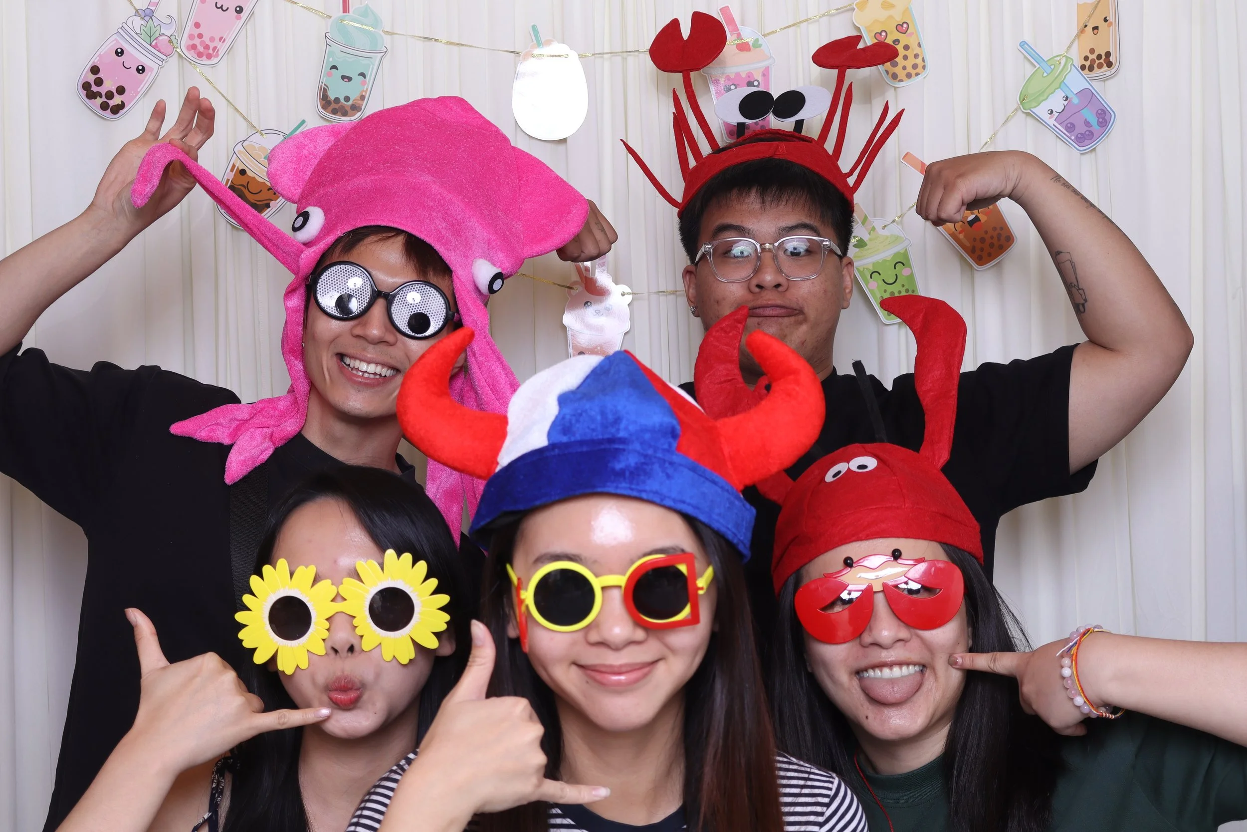 Group of young people wearing playful animal and food-themed hats and sunglasses, celebrating with a cheerful and fun atmosphere, decorated with colorful bubble tea illustrations in the background. Photo Booth Des Moines Iowa