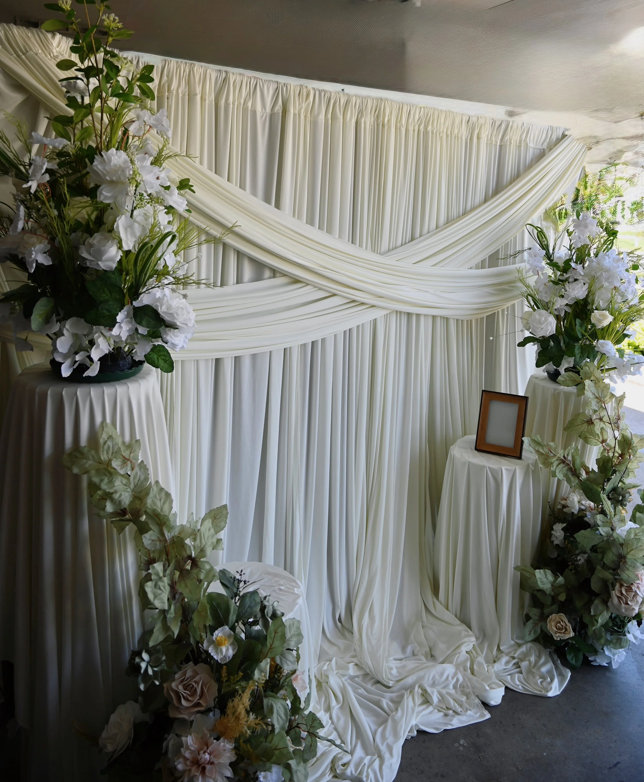 Wedding backdrop with white drapes and floral arrangements on either side, featuring white and blush roses with greenery.