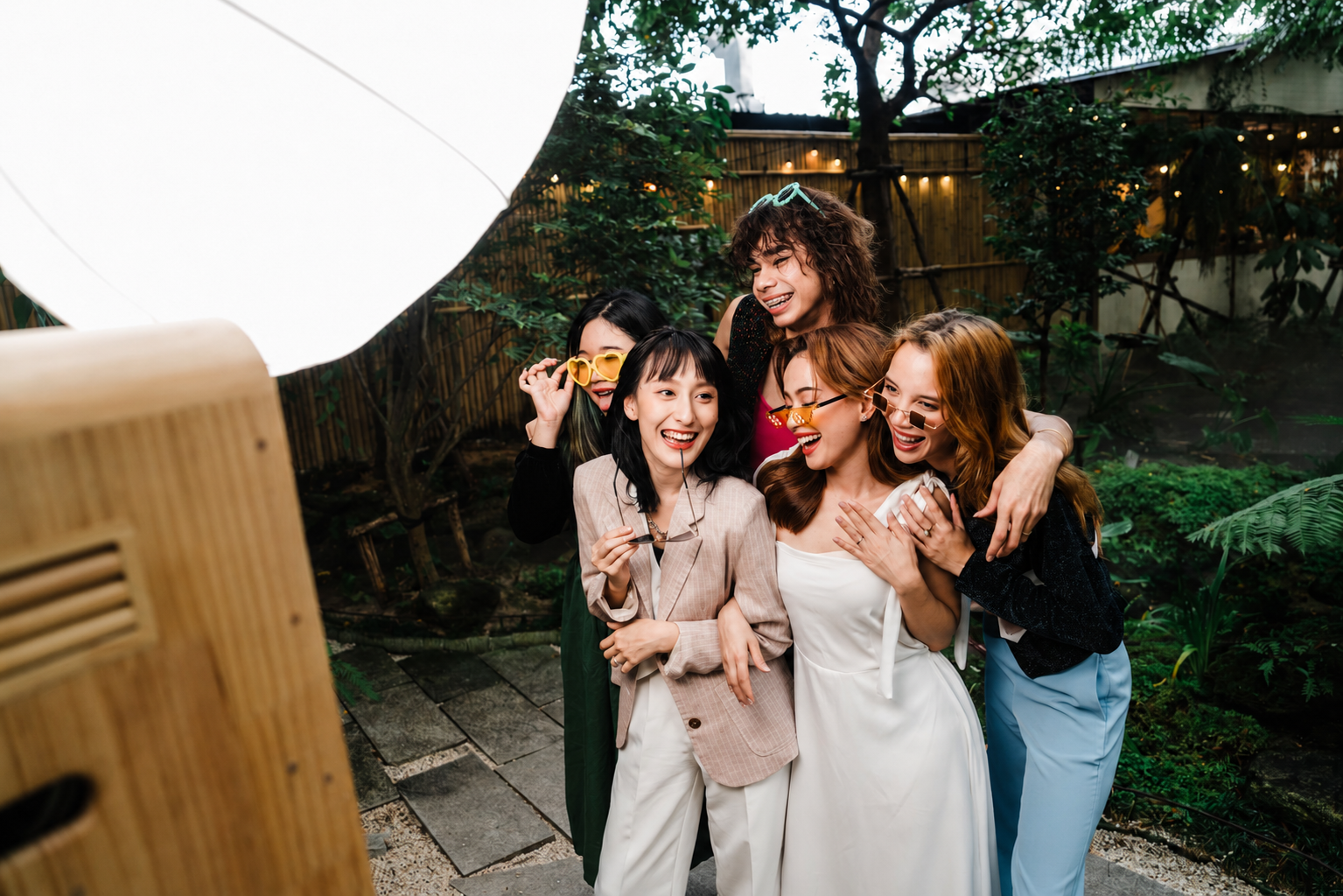 Group of five women celebrating with a photo booth at an outdoor garden party, with one woman in a wedding dress.