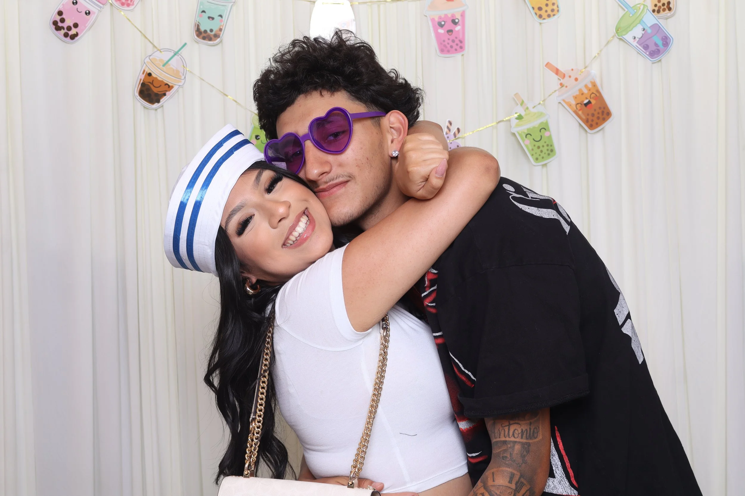 A young woman and a young man are hugging and smiling in front of a colorful bubble tea themed backdrop at a celebration. The woman is wearing a white outfit with a sailor hat and jewelry, while the man is wearing purple heart-shaped sunglasses and a