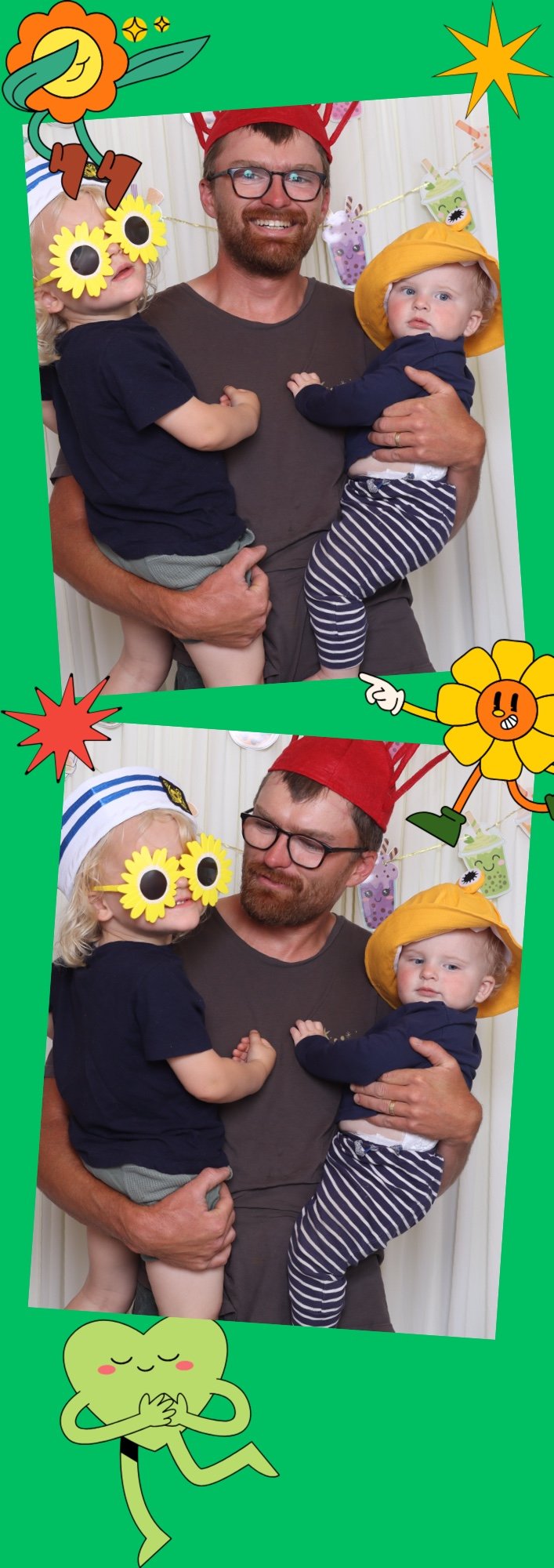 A man with glasses and a beard holding two children, one in each arm, poses in front of a white curtain decorated with colorful party banners. The children wear fun accessories: one has large yellow flower sunglasses and a sailor hat, the other wears