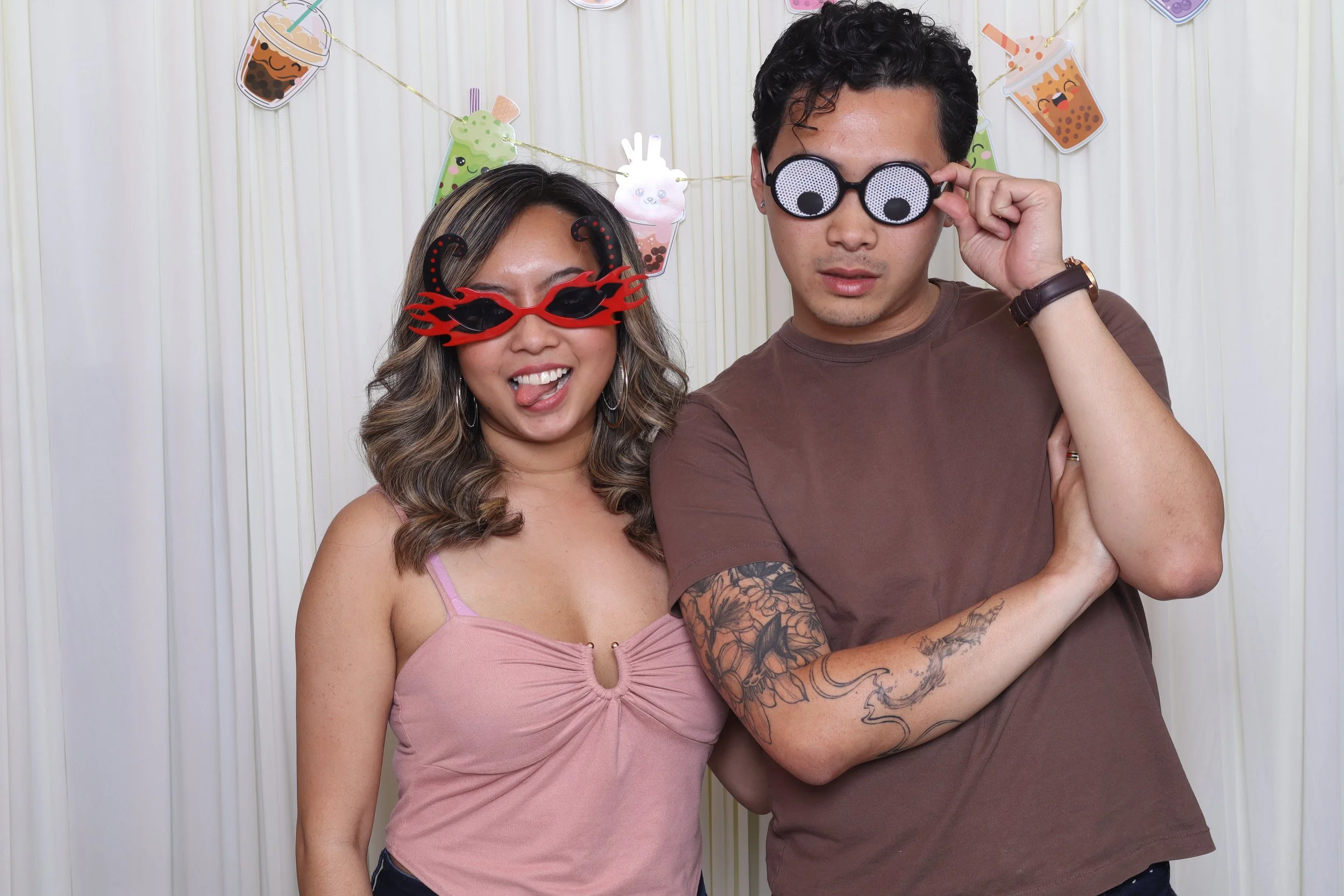 Two people wearing novelty glasses, standing in front of a backdrop decorated with cartoon bubble tea images. The woman on the left wears red glasses with flames design, sticking out her tongue lightly, and has wavy hair with bangs, dressed in a pink