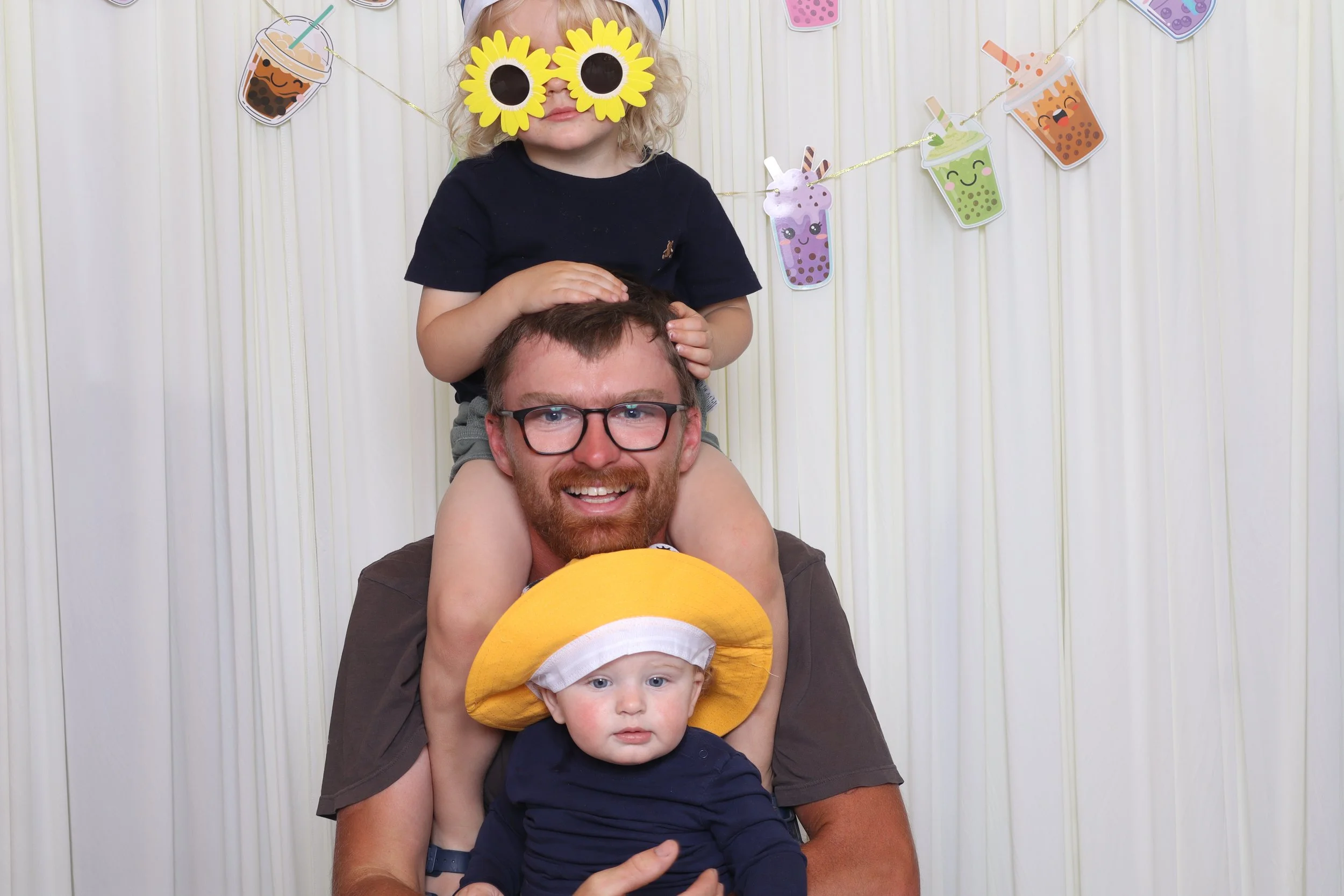 A man with glasses and a beard is sitting with two children on his shoulders against a white curtain background decorated with cartoon bubble tea drink cutouts. The girl on top wears sunflower-shaped sunglasses and a black shirt, while the boy below 