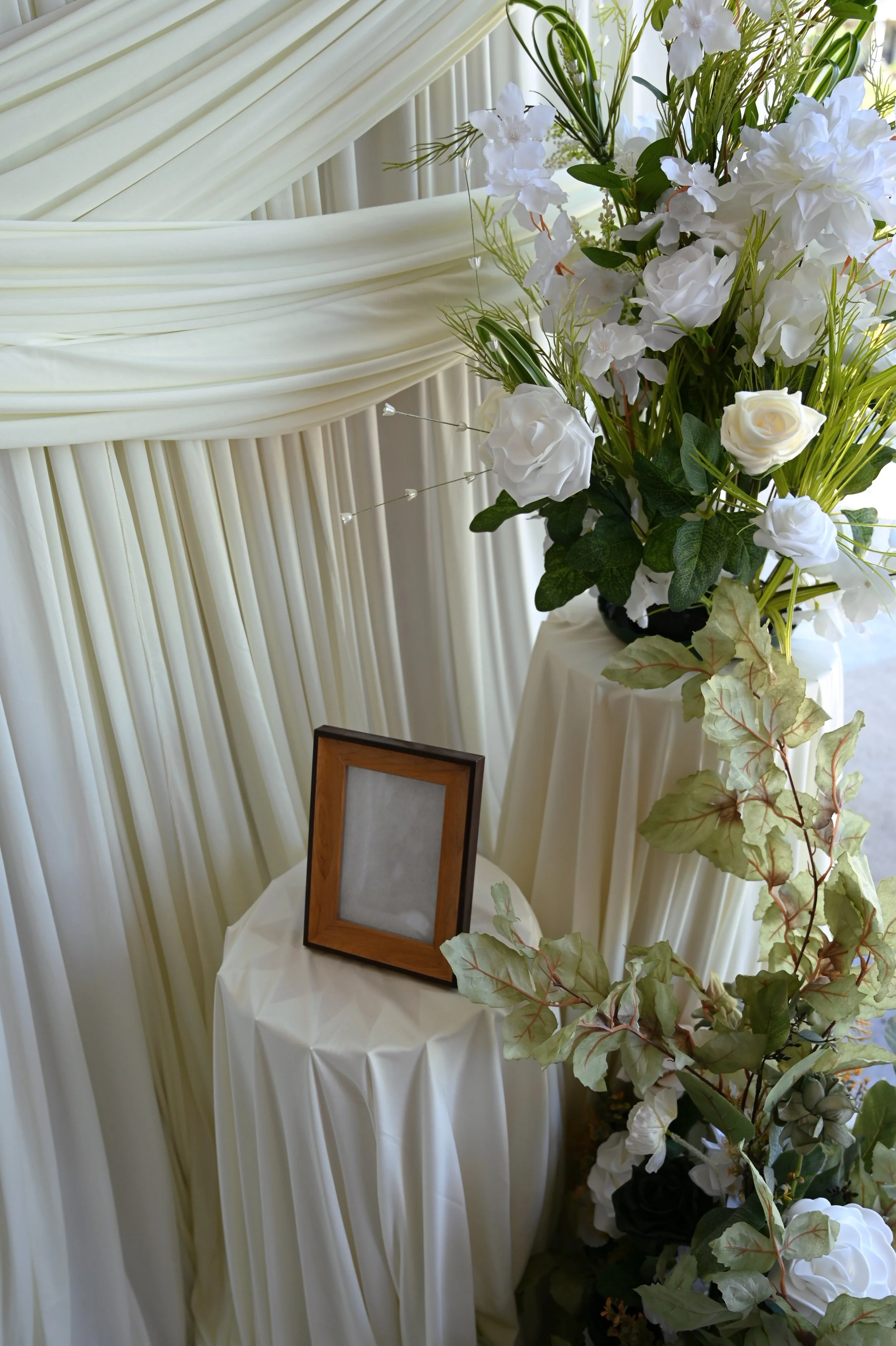 Sample of a floral arrangement with white flowers, green leaves, and draped white fabric in the background, possibly at a wedding or event setting.