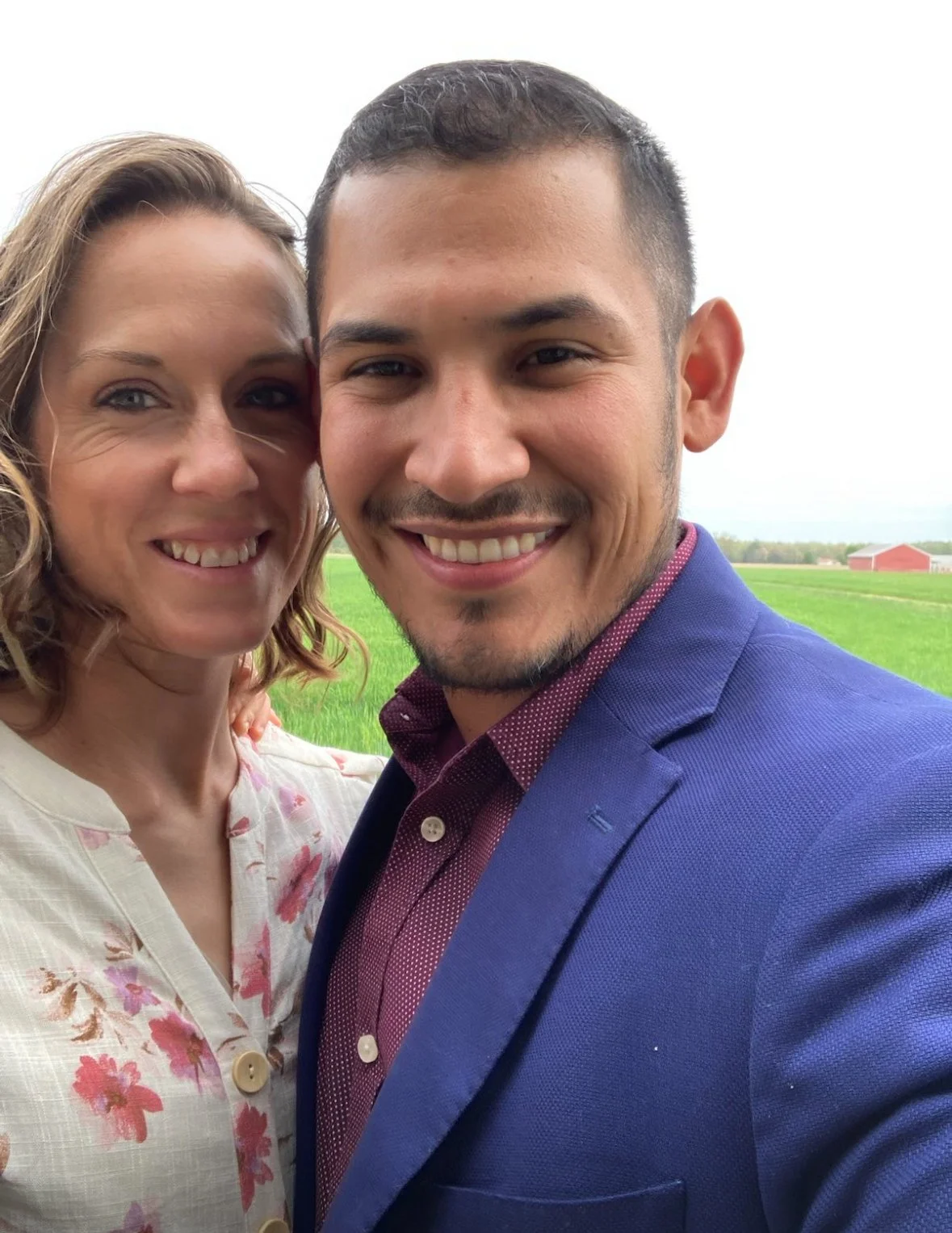 A smiling woman and man taking a selfie outdoors in a green field with a red barn in the background.