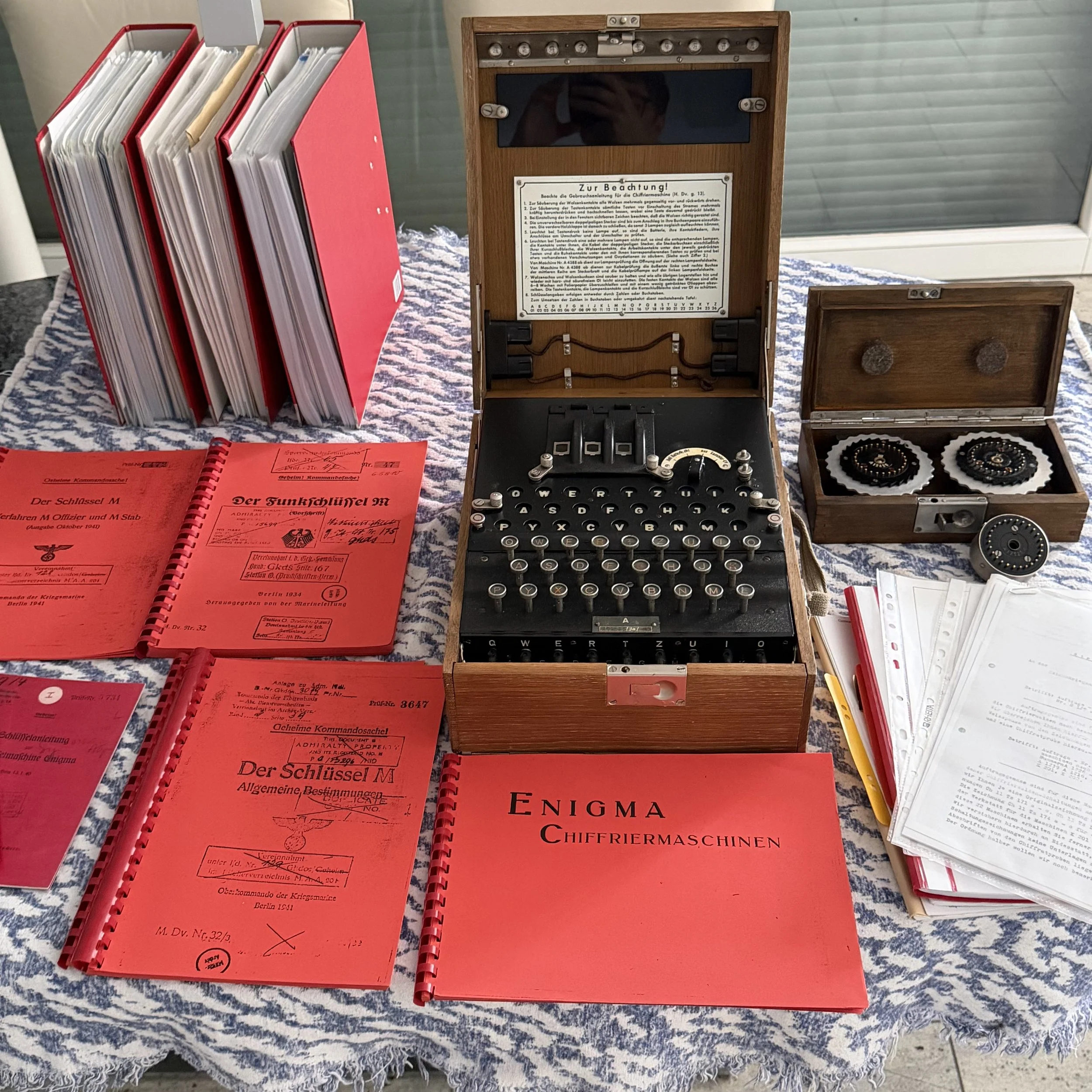 Collection of red and white paper documents, a wooden cipher enigma machine, and various booklets and papers on a textured tablecloth.