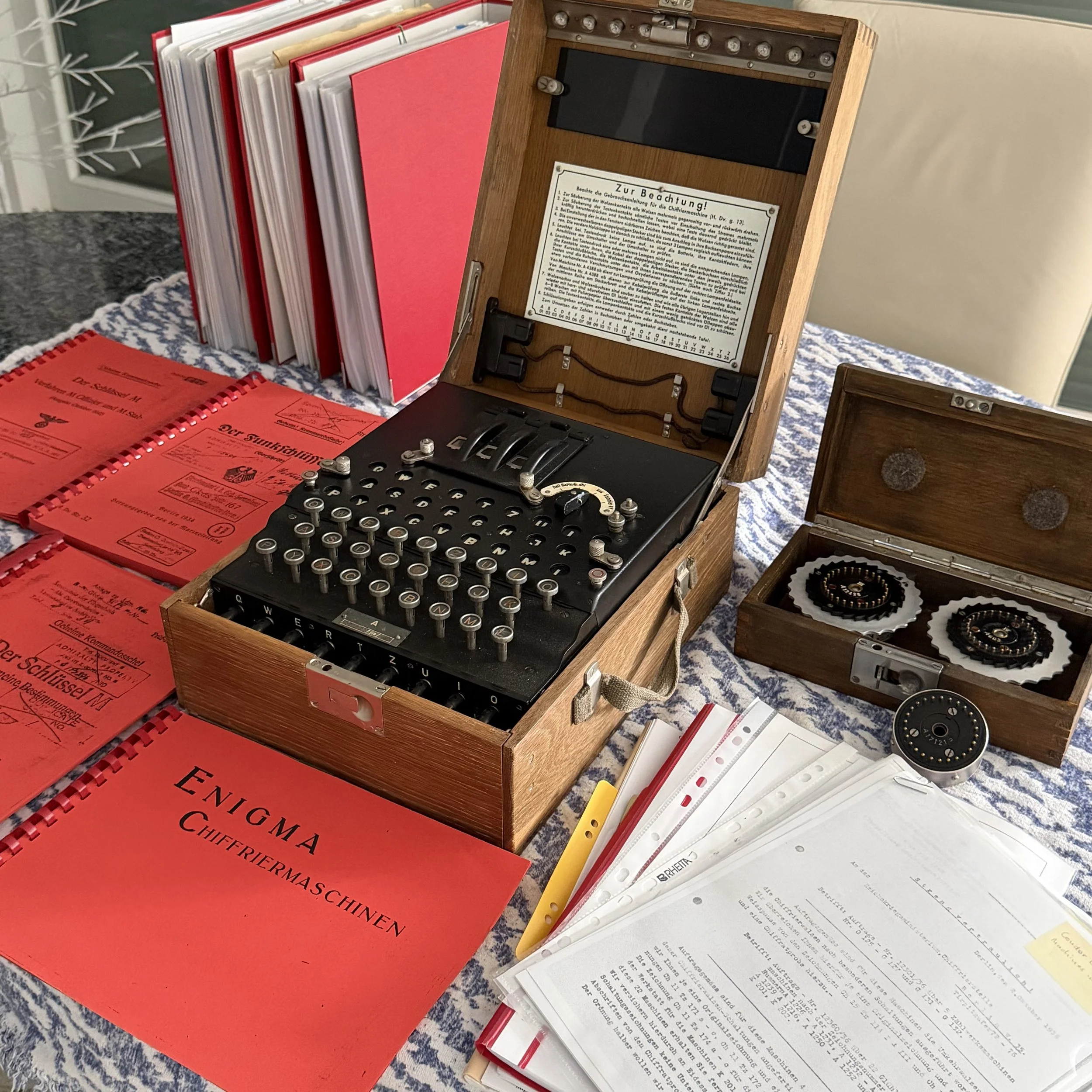 A vintage mechanical typewriter in a wooden case with sheet music and music folders around it. The scene appears to be on a table with a patterned tablecloth, and books or documents are also present.