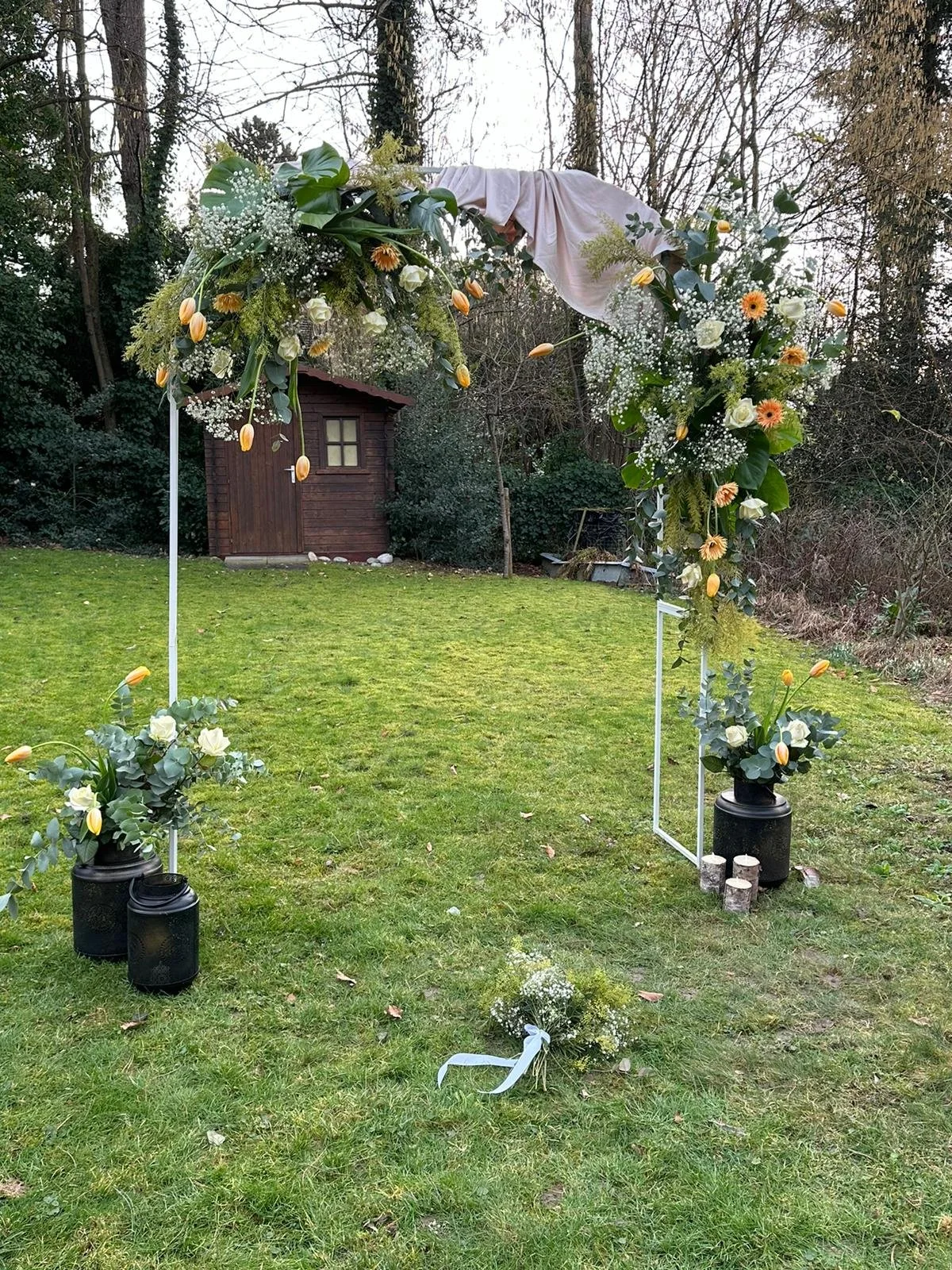 Blumenbogen für eine Hochzeit in einem Garten mit grüner Wiese, Holzhütte im Hintergrund, und Blumenarrangements aus weißen Rosen, Tulpen und anderem mit Schleifen und Vasen