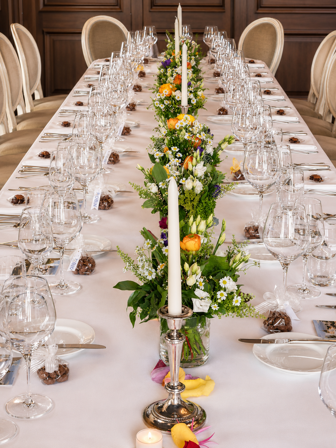 Es ist ein gedeckter Festtafeltisch mit Blumenarrangement in der Mitte, Kerzen, Weingläser, Besteck und kleine Geschenke unter weißen Tischdecken.