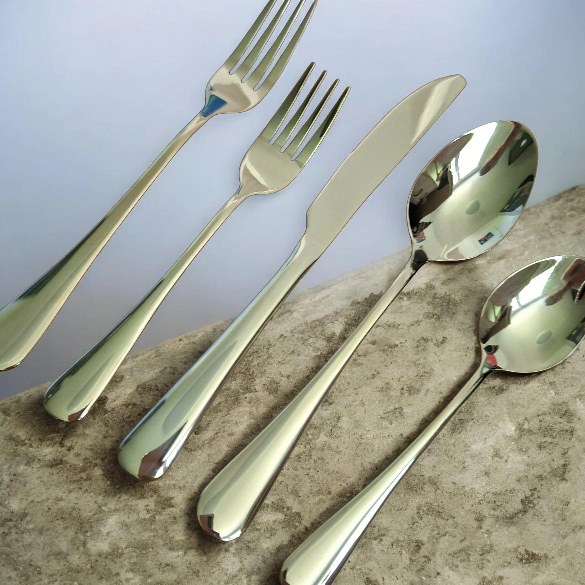 Mehrere goldene Besteckteile, darunter drei Gabeln, eine Messerklinge und ein Löffel, liegen auf einer grauen Oberfläche. Das Besteck hat eine glänzende Oberfläche, die die Umgebung reflektiert.