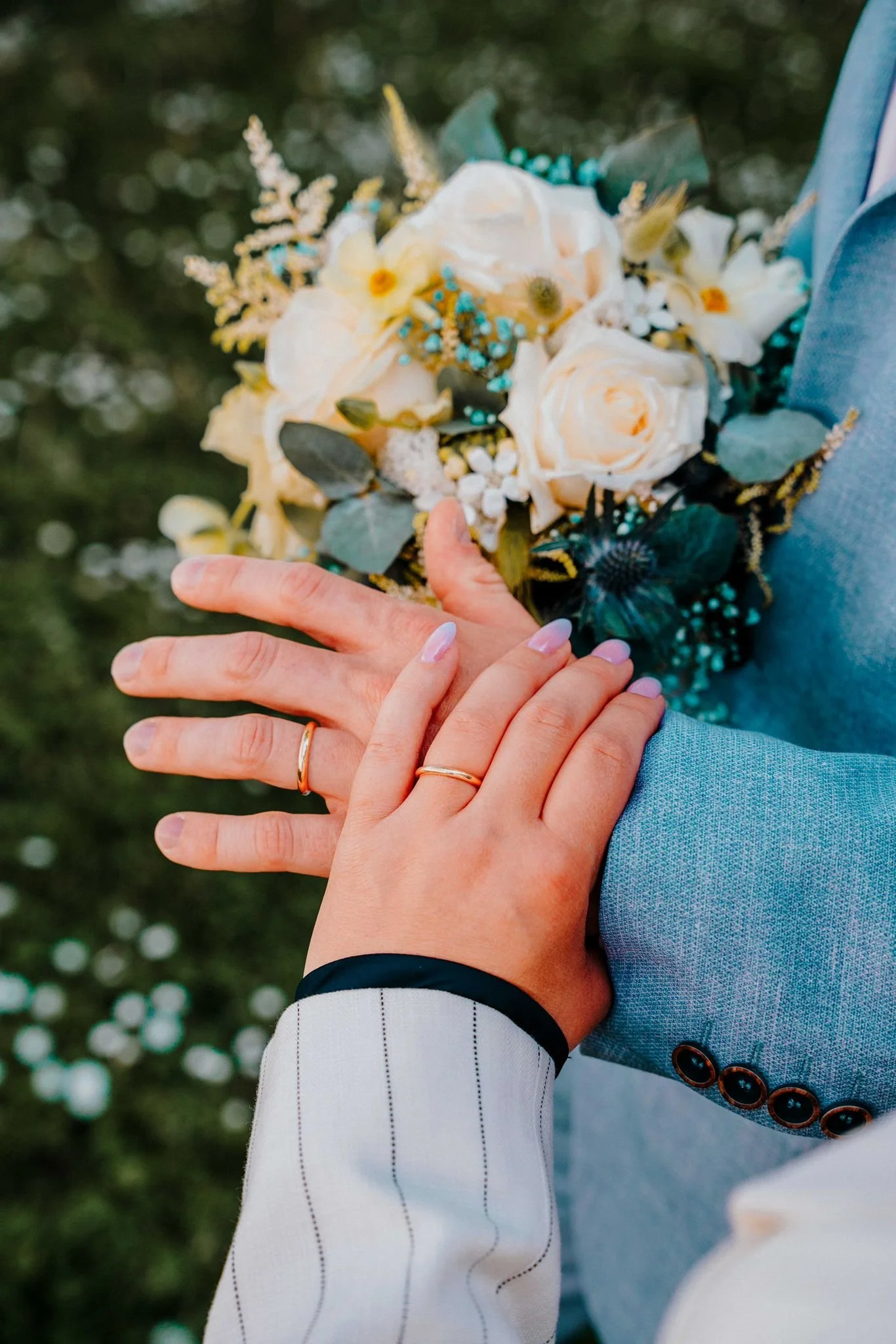 Ein Paar hält sich an den Händen, wobei die linke Hand einer Frau mit Nagellack und Ehering sichtbar ist, während die rechte Hand eines Mannes sichtbar ist. Der Mann trägt einen hellblauen Anzug mit braunen Knöpfen, die Frau trägt einen weißen Blazer mit schwarzen Nadelstreifen. Im Hintergrund sind Blumen und ein unscharfer grüner Garten zu sehen