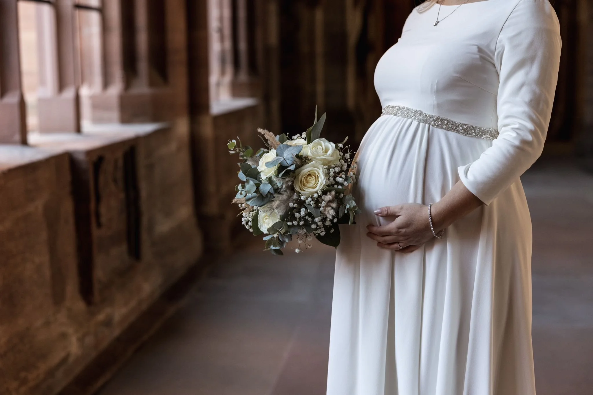 Eine Frau in einem weißen Brautkleid hält einen Brautstrauß aus weißen Rosen und verschiedenen Grünpflanzen in den Händen.