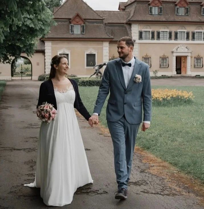 Ein Brautpaar, das Hand in Hand spazieren geht, vor einem historischen Gebäude, die Braut trägt ein weißes Kleid und hält einen Blumenstrauß, der Bräutigam trägt einen blauen Anzug mit Fliege.
