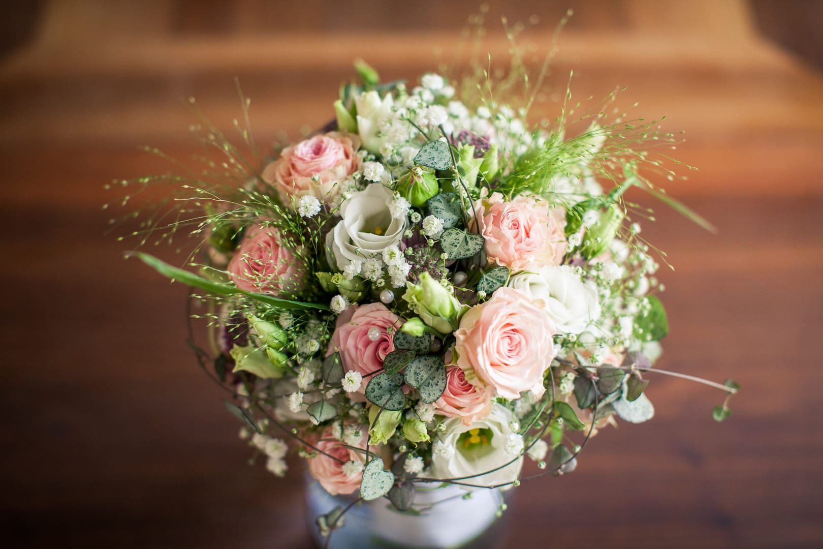 Blumenstrauß mit rosa Rosen, weißen Calla und kleinen weißen Blüten in einer Vase auf Holzfläche.