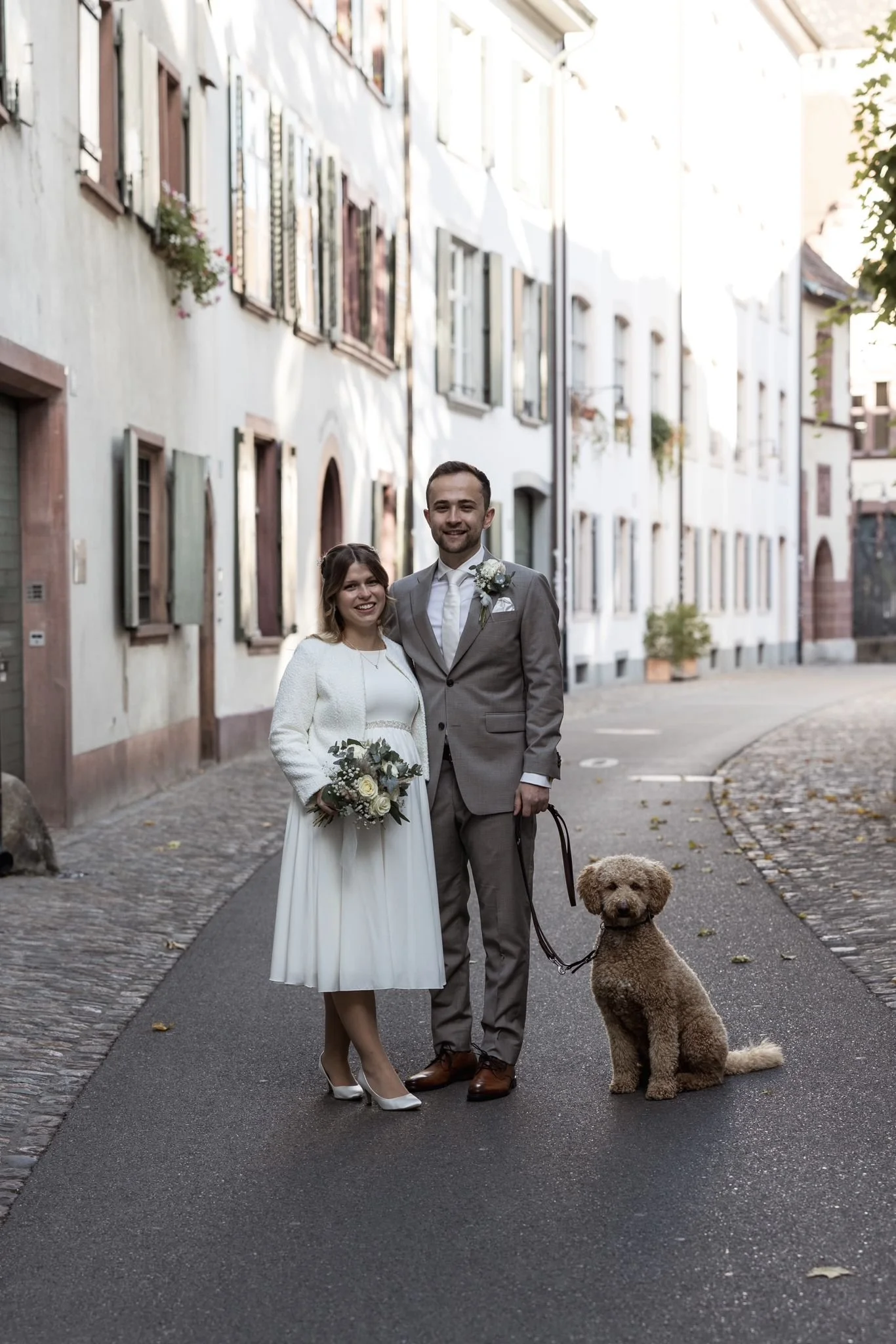 Ein Brautpaar steht auf einer Kopfsteinpflasterstraße, die Weg wird gesäumt von alten Häusern. Die Braut trägt ein langes weißes Kleid mit einem Blazer und hält einen Brautstrauß. Der Bräutigam trägt einen grauen Anzug mit Krawatte und hat einen Blumenanstecker am Revers. Ein brauner Hund sitzt an der Leine des Mannes.