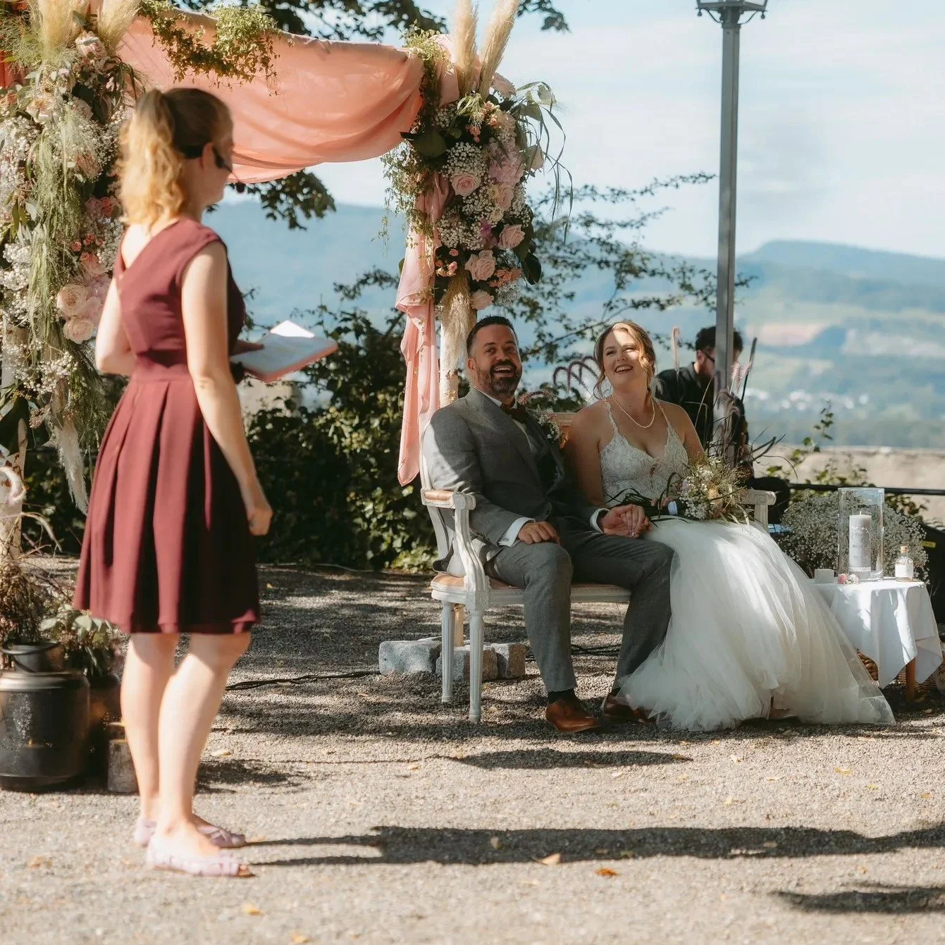 Ein Brautpaar sitzt auf einer Bank, während eine Frau in einem roten Kleid eine Rede hält. Das Brautpaar lächelt glücklich, im Hintergrund ist eine schöne Landschaft mit Bergen zu sehen. Es ist eine Hochzeitsfeier im Freien mit Blumen und Dekorationen.