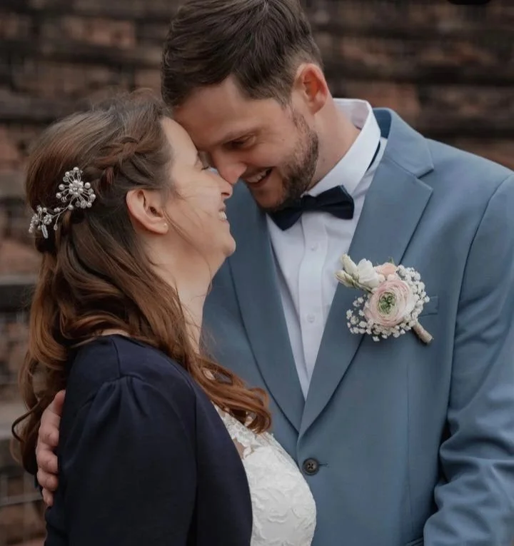 Ein glückliches Brautpaar bei einer Hochzeit, blickt sich liebevoll ins Gesicht, Händchen haltend, vor einer holzvertäfelten Wand.