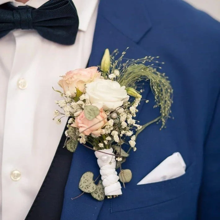An einem dunklen blauen Anzug mit weißem Hemd und schwarzer Fliege befestigt sich eine Brautjungfernsbouquet aus weißen und rosa Blumen, Federn und grünem Blatt.