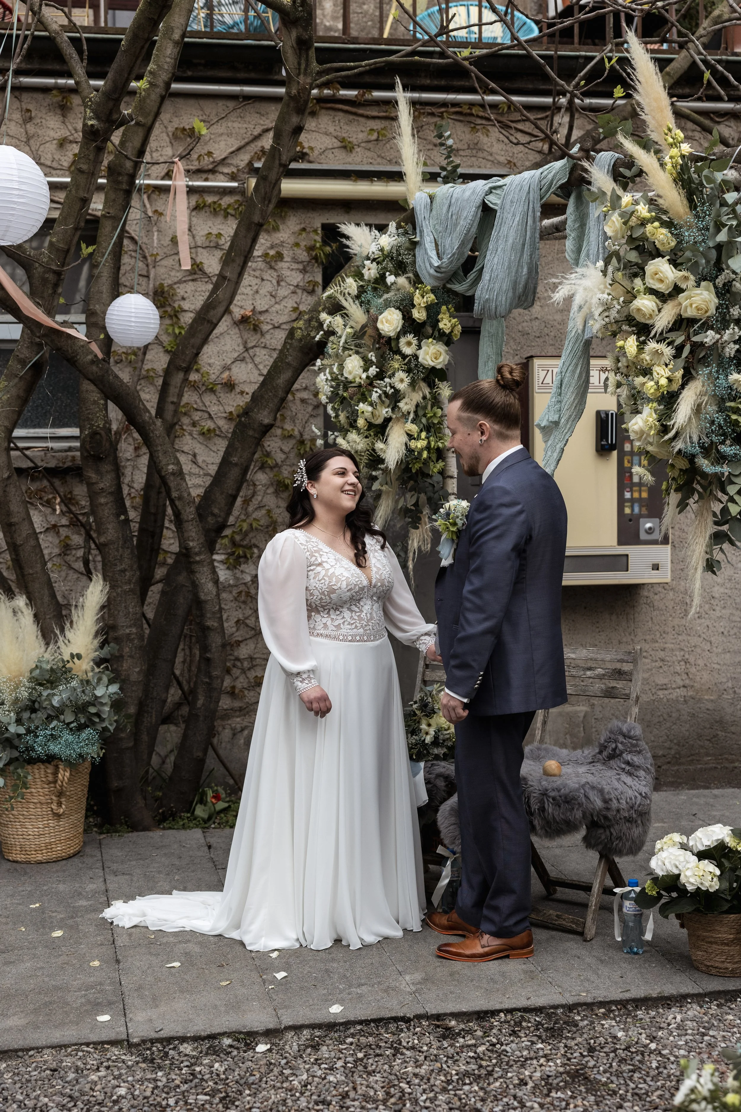 Ein Hochzeitspaar steht sich gegenüber unter einem Blumenbogen im Garten. Die Braut trägt ein weißes Hochzeitskleid, der Bräutigam einen dunkelblauen Anzug. Im Hintergrund sind weiße Blumen und Dekorationen, sowie Gartenelemente sichtbar.