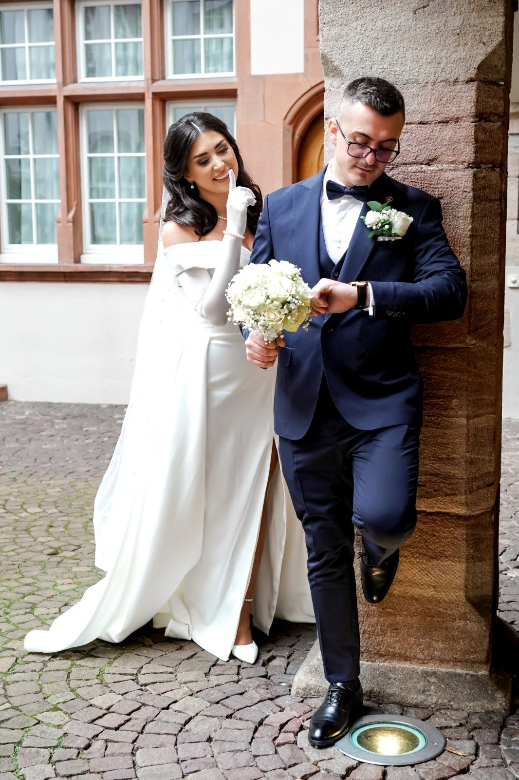 Ein Brautpaar bei ihrer Hochzeit, die Frau im weißen Brautkleid, der Mann im dunklen Anzug, sie stehen außerhalb vor einer alten Steinswand, der Mann schaut auf seine Uhr, die Braut hält einen Blumenstrauß.