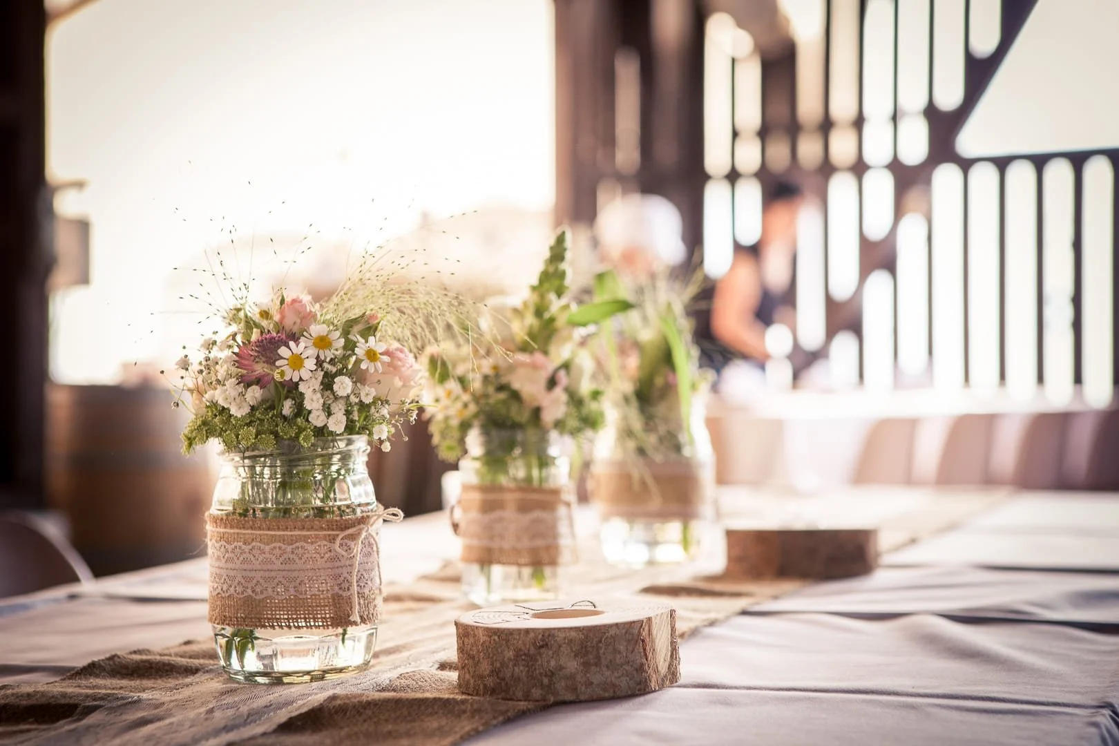 Blumenarrangement auf einem Tisch mit Holzscheiben und Stoffabschirmungen, im Hintergrund eine helle, sonnige Innenraumumgebung.