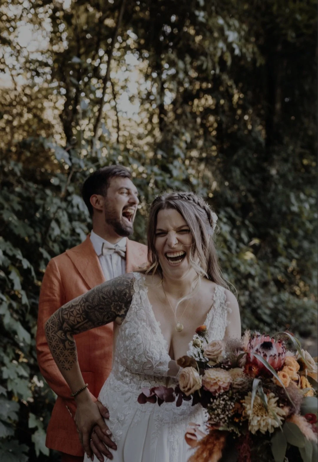 Ein lachendes Paar bei einer Hochzeit im Freien, umgeben von Bäumen, das Brautkleid mit Blumen und Tattoos zeigt.