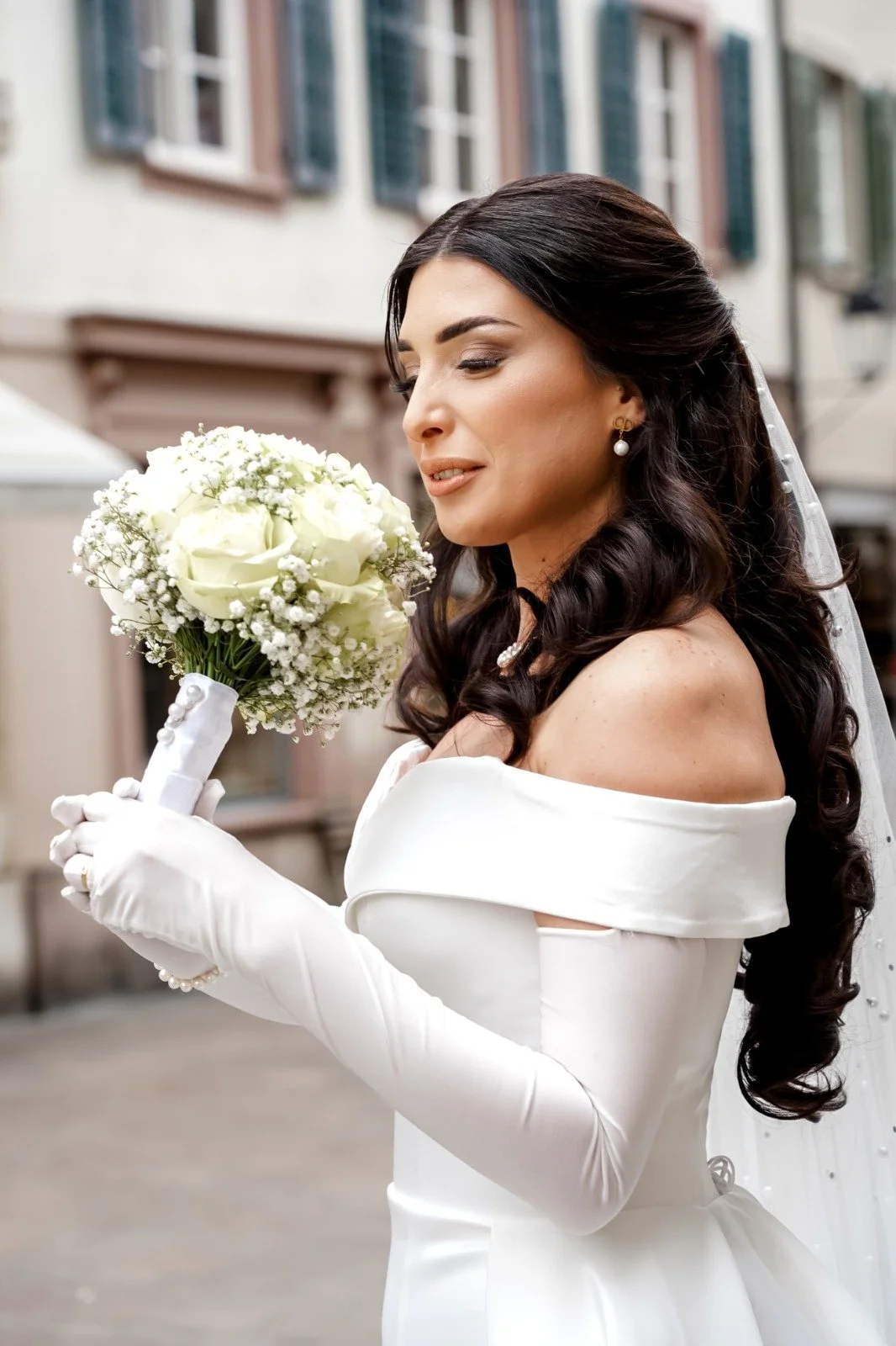 Eine Braut in einem weißen Kleid mit schwarzen Haaren hält einen Brautstrauß aus weißen Rosen und kleinen weißen Blümchen, steht vor einem Mehrfamilienhaus mit offenen Fenstern.