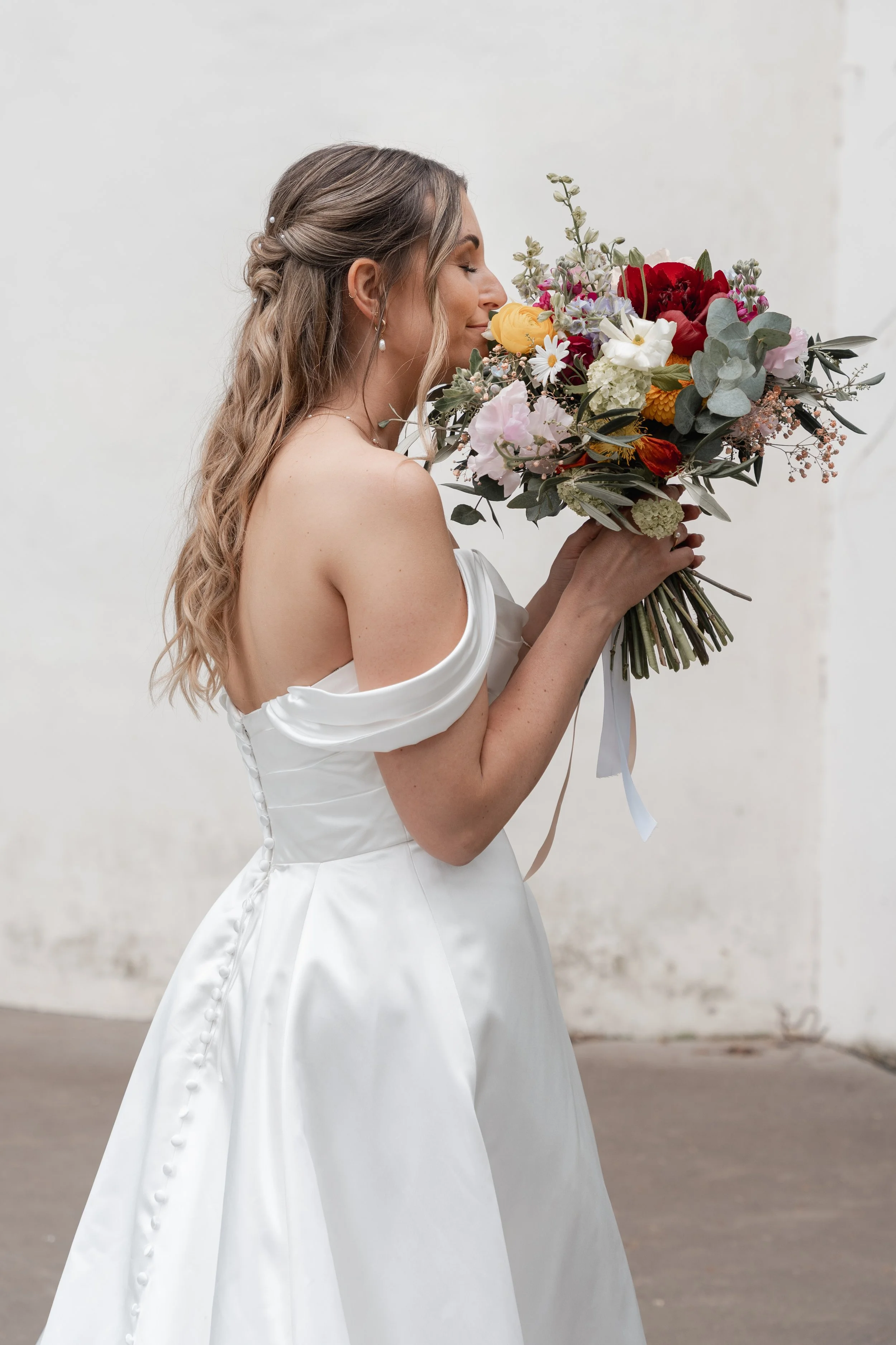Eine Frau in einem weißen Brautkleid hält einen Blumenstrauß, hat gelocktes, langes Haar mit kleinen Akzenten, trägt Perlenohrringe, steht vor einer weißen Wand.