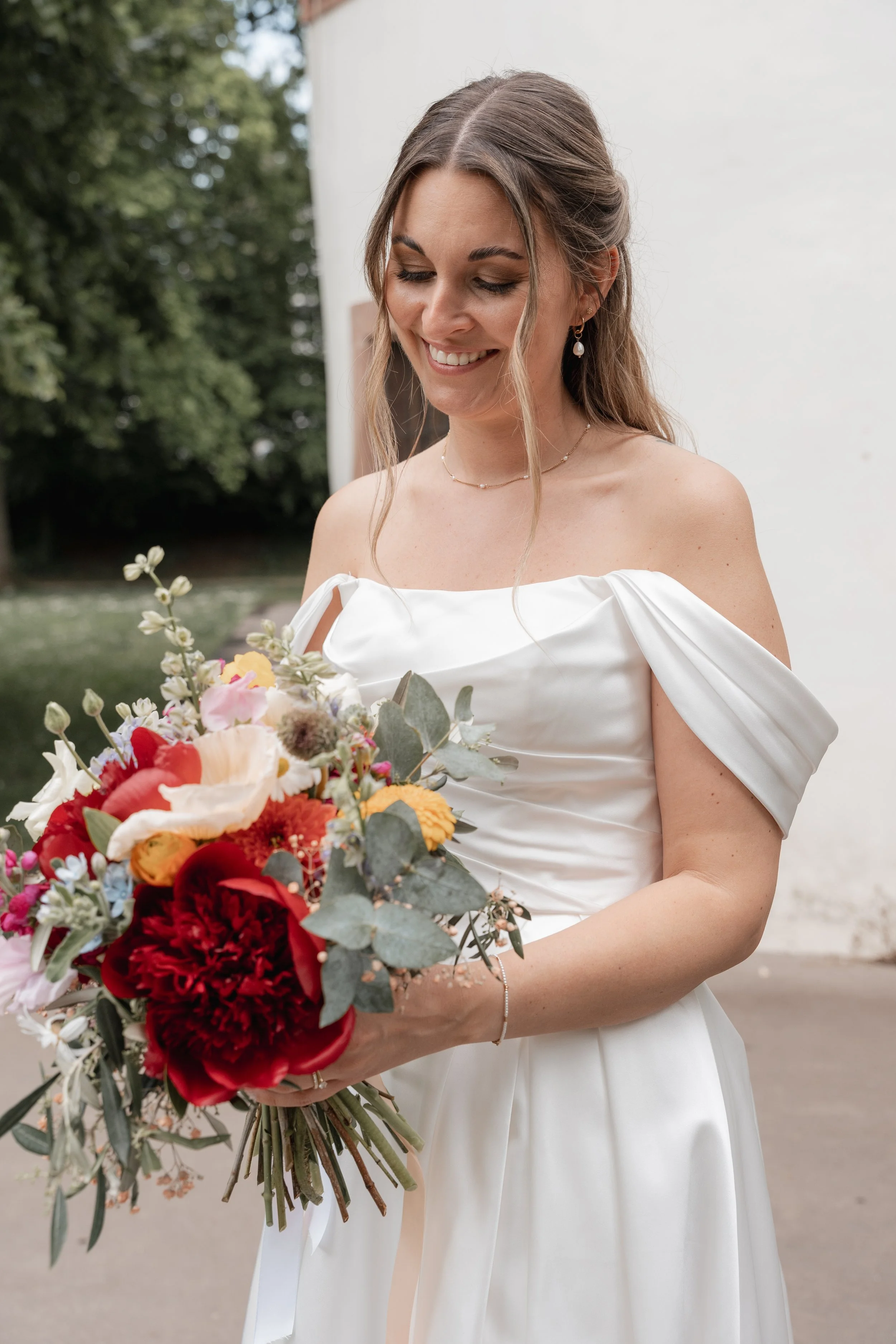 Eine lachende Frau in einem weißen Brautkleid hält einen bunten Blumenstrauß bei einer Hochzeit im Freien.