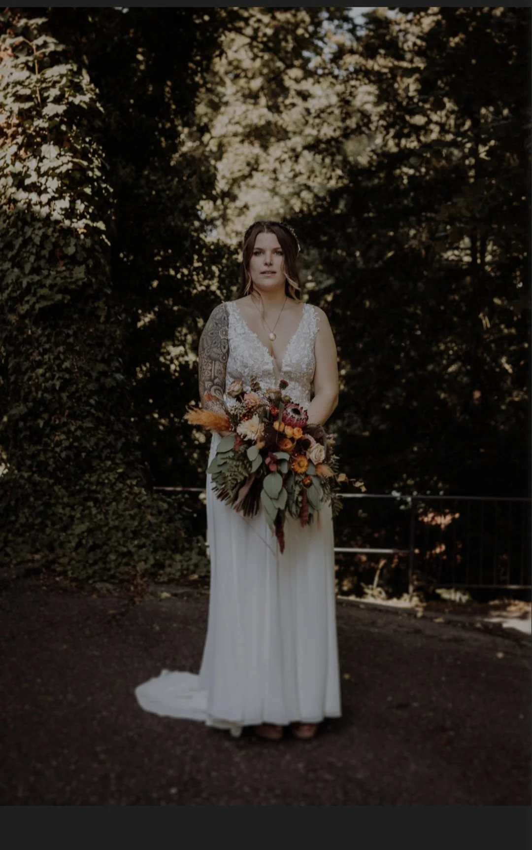 Eine Frau in einem weißen Brautkleid hält einen großen Blumenstrauß und steht vor einem dunklen, bewaldeten Hintergrund.