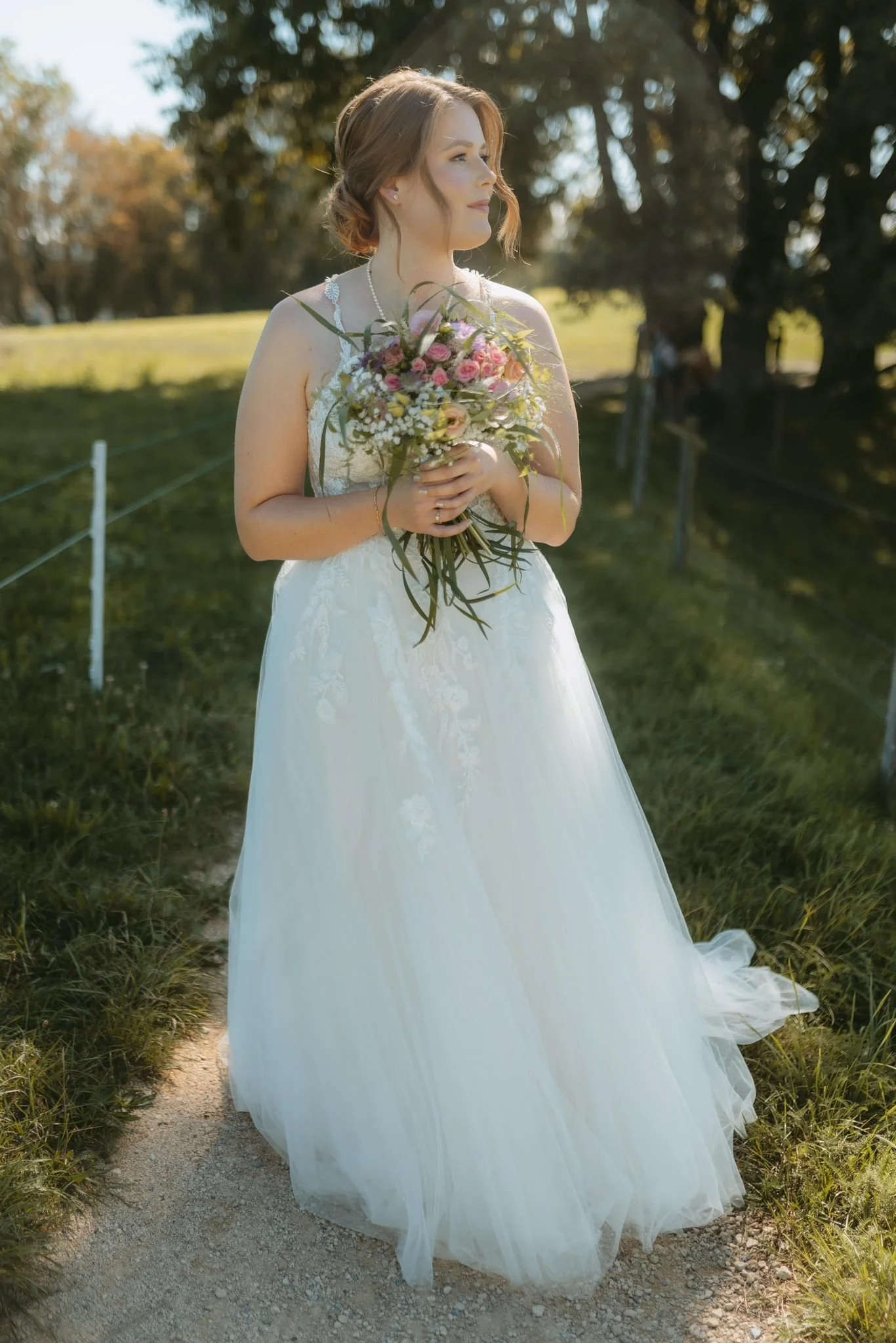 Eine Frau in einem weißen Brautkleid hält einen Blumenstrauß bei Sonnenuntergang im Freien, umgeben von Bäumen und Rasen.