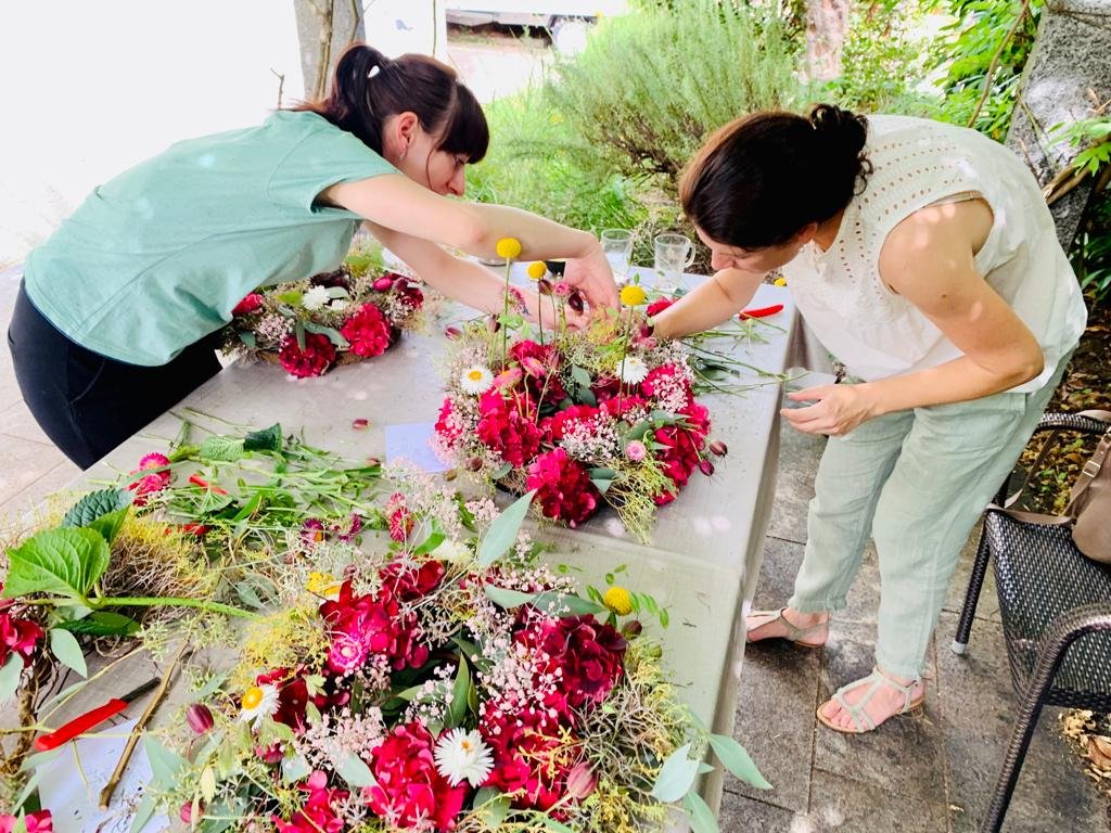 Zwei Frauen arbeiten gemeinsam an Blumengestecken auf einem Tisch. Der Tisch ist voller verschiedener Blumen und Pflanzen, die sie arrangieren.