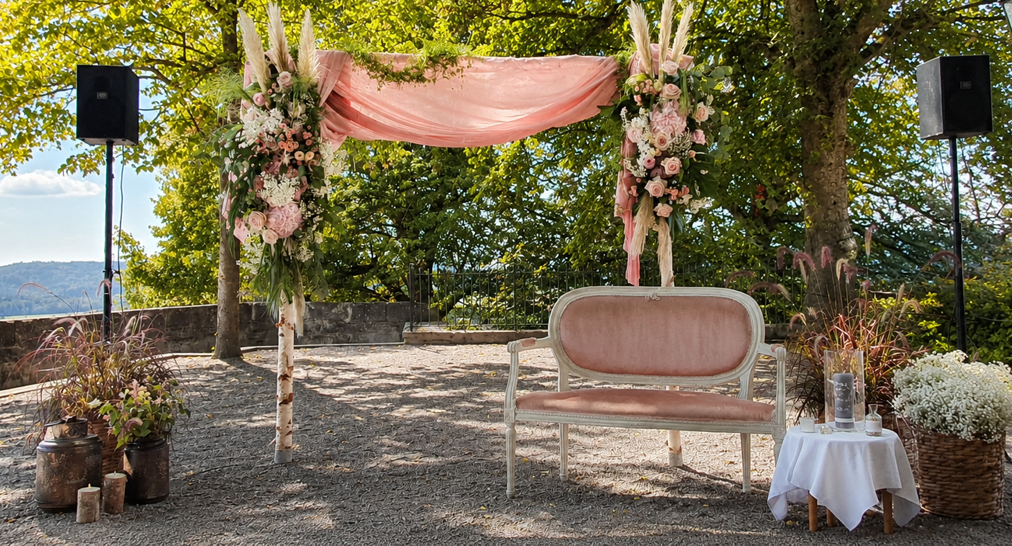 Ein festlich dekorierter Bereich im Freien mit einem pinken, stilvollen Sofa vor einem Blumenbogen aus rosa und weißen Blumen, umgeben von Pflanzen, Kerzen und einem kleinen Tisch mit weißen Blumen und Gegenständen darauf, umgeben von Bäumen und einer Landschaft im Hintergrund.