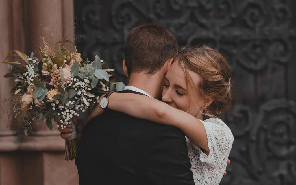 Braut und Bräutigam umarmen sich liebevoll bei ihrer Hochzeit, die Braut hält einen Blumenstrauß, im Hintergrund eine dunkle ornamente Wand.