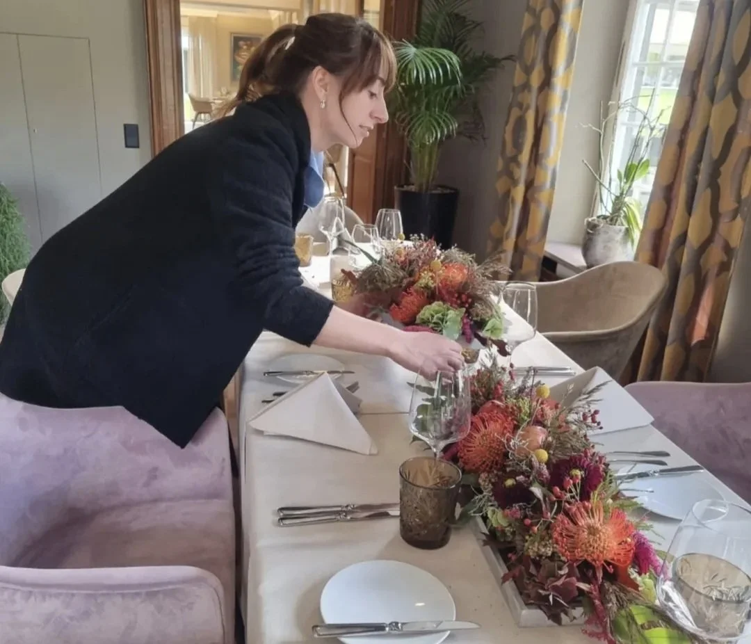 Frau richtet einen festlich gedeckten Tisch mit Blumenarrangements, Weingläsern, Tellern, Besteck und Kerzen in einem dining Raum mit großen Fenstern und Dekorationen.