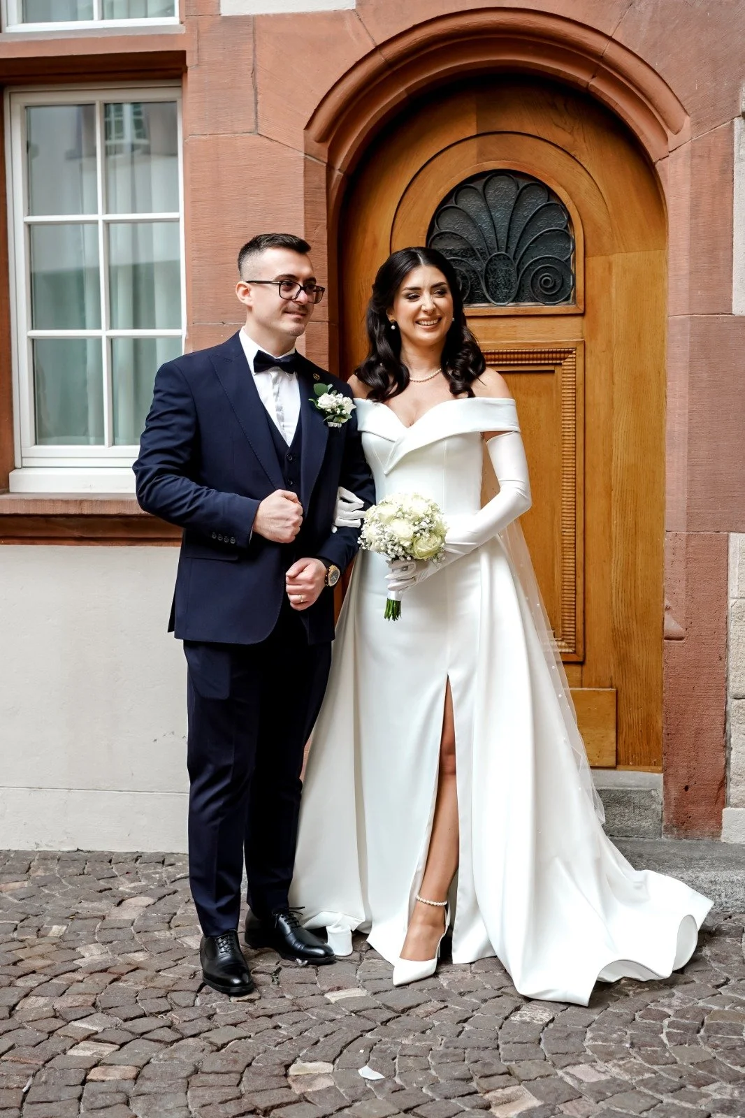Ein frisch verheiratetes Paar, der Bräutigam in Anzug und Fliege, die Braut in einem weißen Hochzeitskleid mit Beinschlitz, steht vor einem Haus mit roten Ziegelsteinen und Holz-Tür.