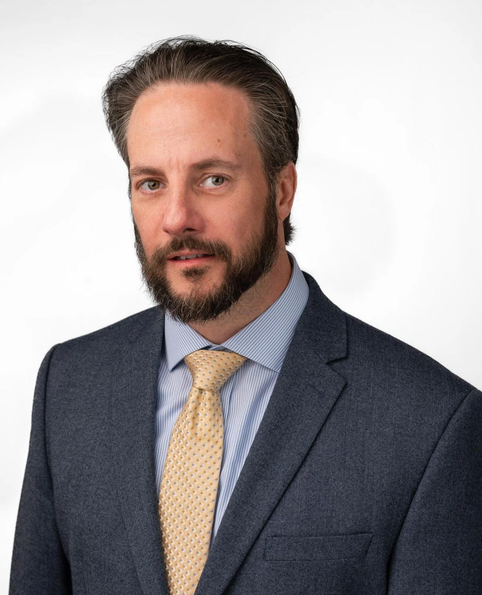 Close-up of a man with dark hair and a beard, wearing a suit with a yellow tie, standing against a plain white background.