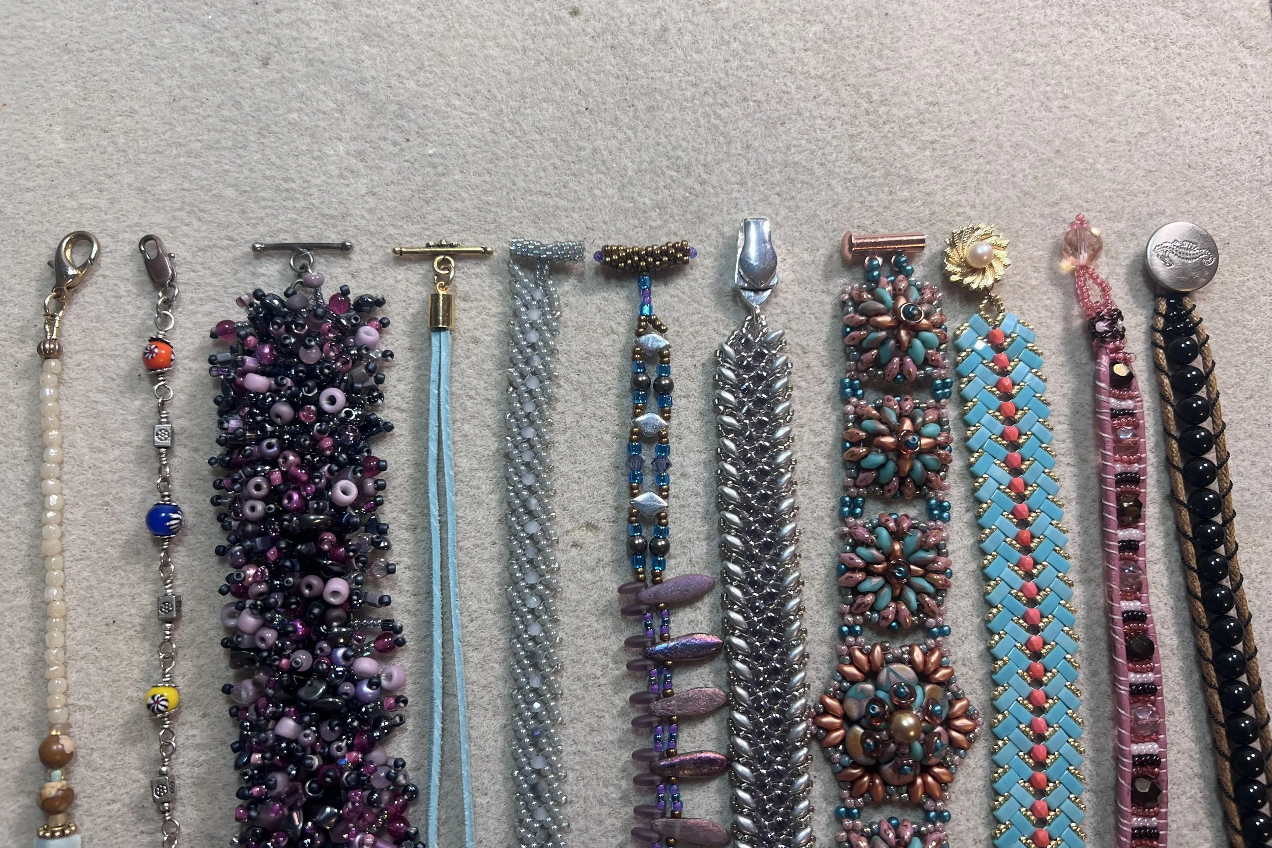 A lineup of handmade jewelry closures showing two styles of lobster clasps, a beadwoven toggle, a leather crimp-and-toggle closure, handmade beaded toggles, a magnetic clasp, a barrel clasp, a box clasp, and a beaded loop-and-button closure.