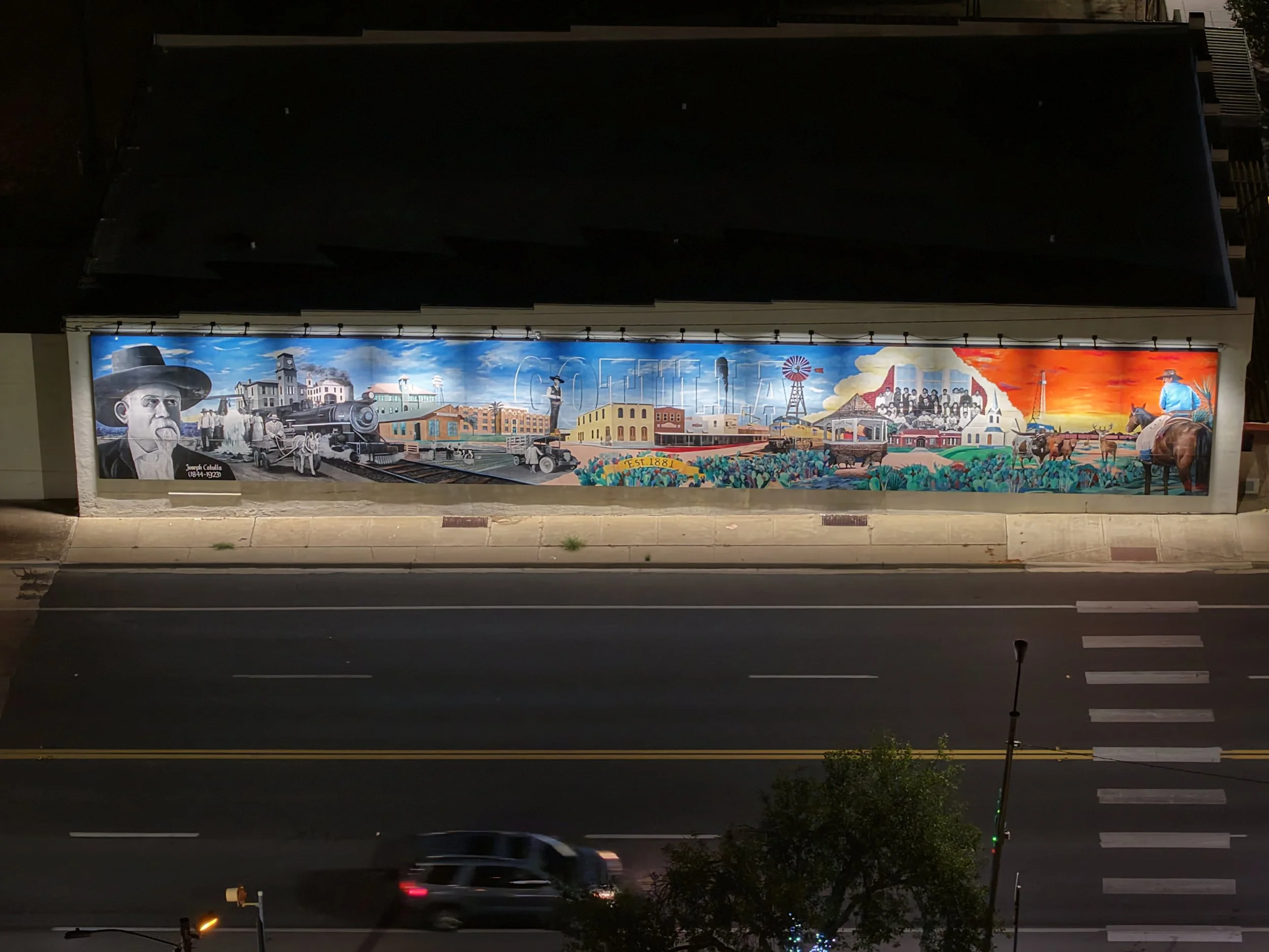 Nighttime mural on a building depicting historical scenes, including a man in a hat, a train, a Carousel, wind turbines, and a cowboy on a horse.