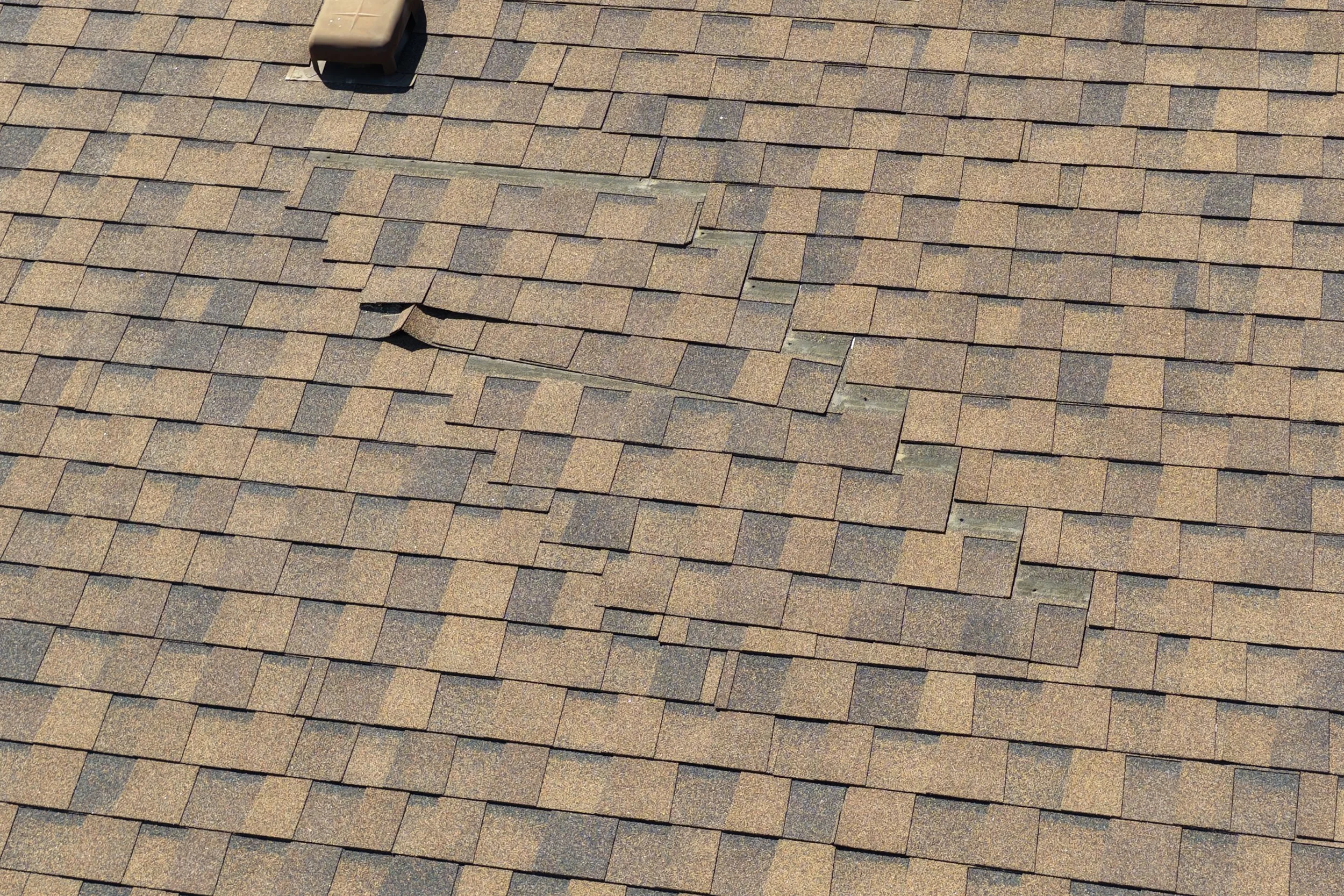 Close-up view of a roof covered with brown shingles, showing a small section with some shingles slightly displaced and a vent pipe in the upper left corner.