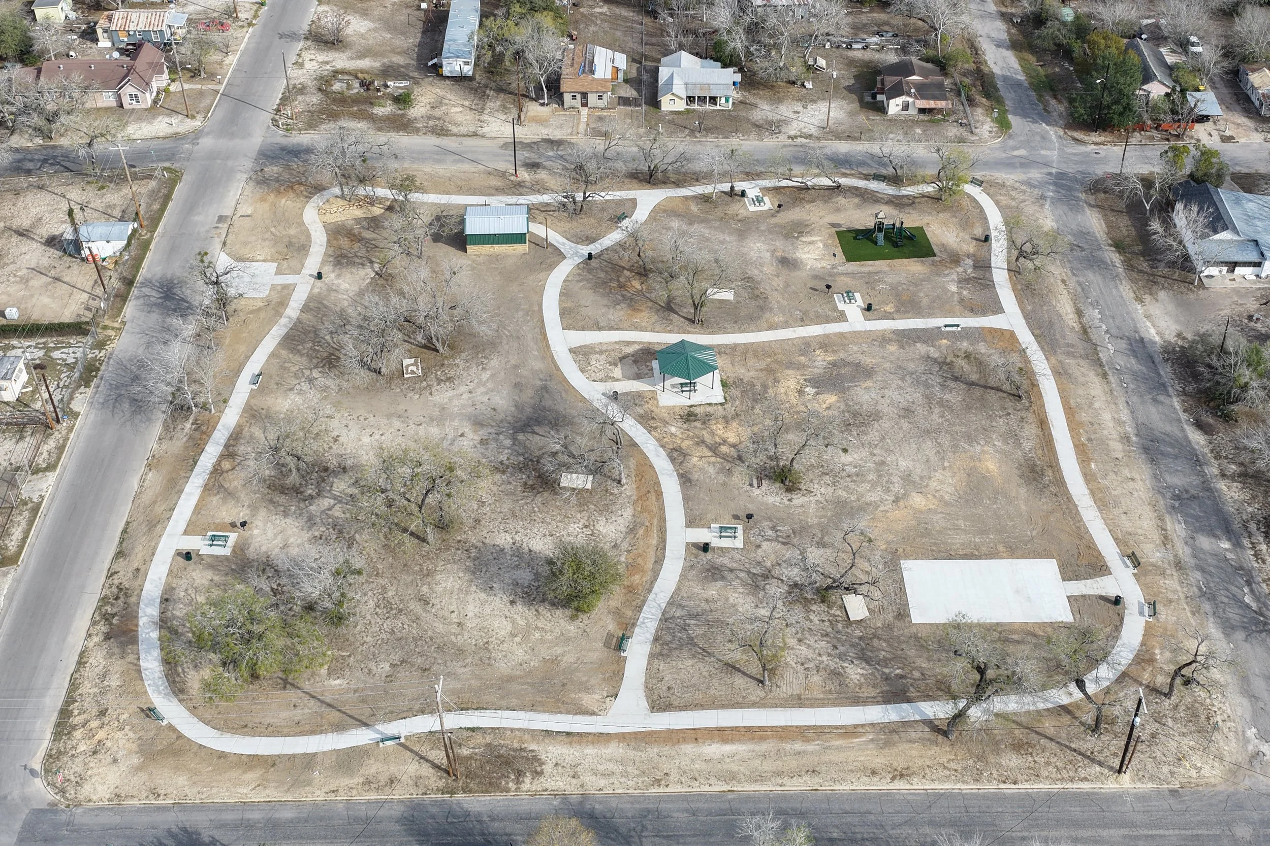 Aerial view of a small park with winding concrete pathways, a green gazebo, trees, benches, trash cans, and a playground area on a partly cloudy day in an urban neighborhood with houses and roads surrounding the park.