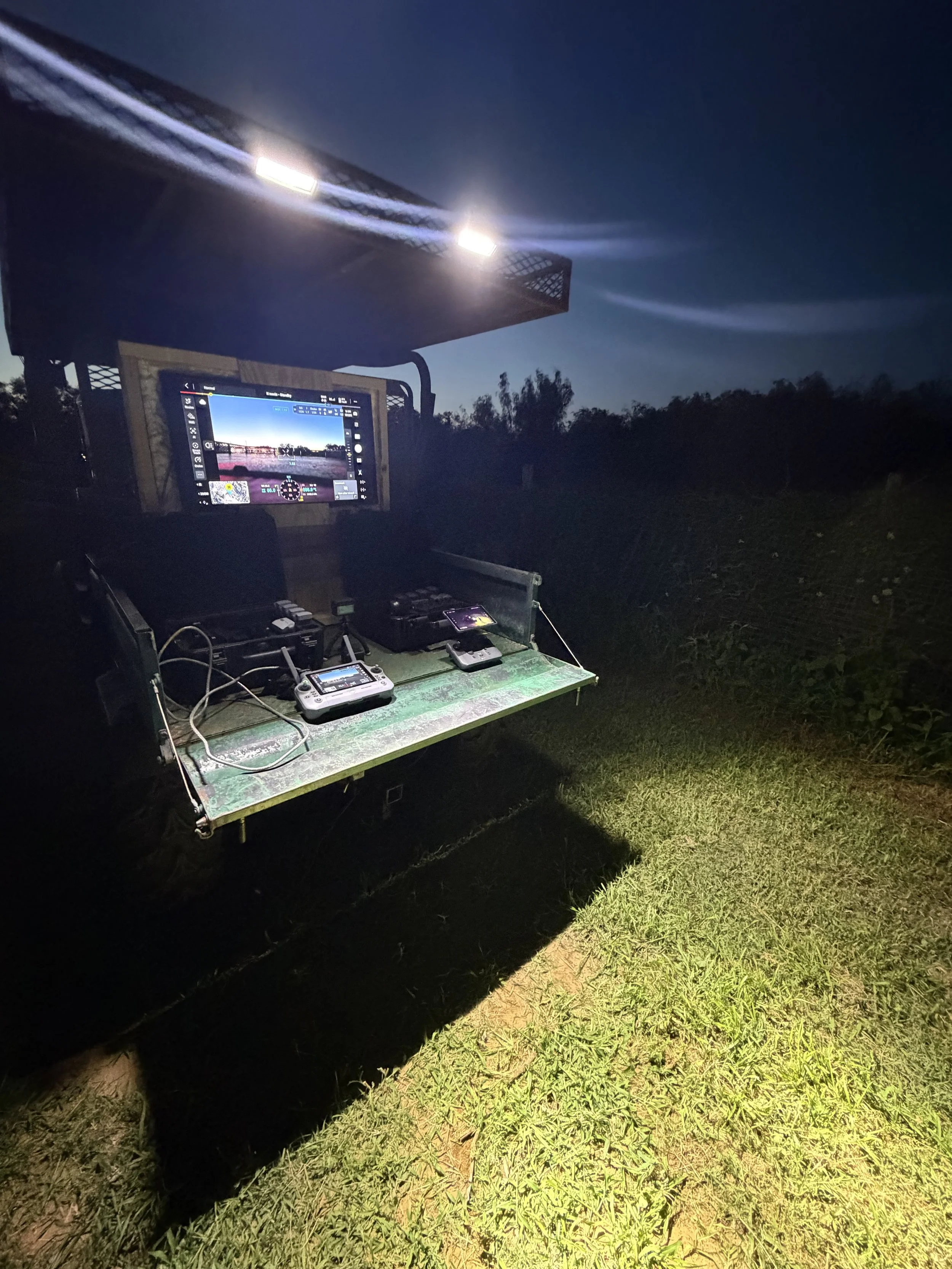 Drone control station with a monitor displaying a drone flight at night, set up outdoors on a green table with various equipment, illuminated by two bright lights in the dark.