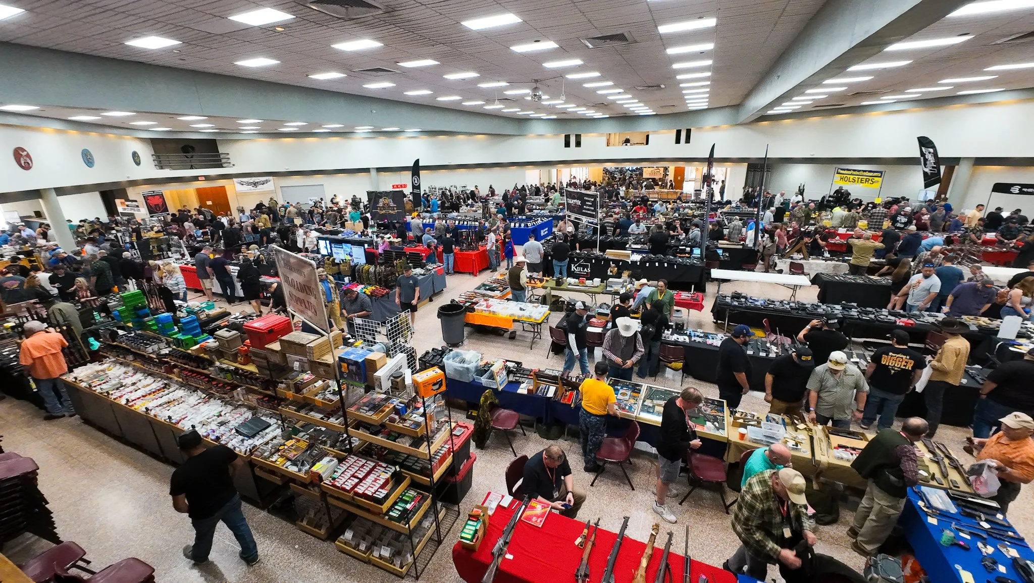 A large indoor marketplace with many booths selling various items, filled with shoppers and vendors. The space has a high ceiling with bright overhead lighting.