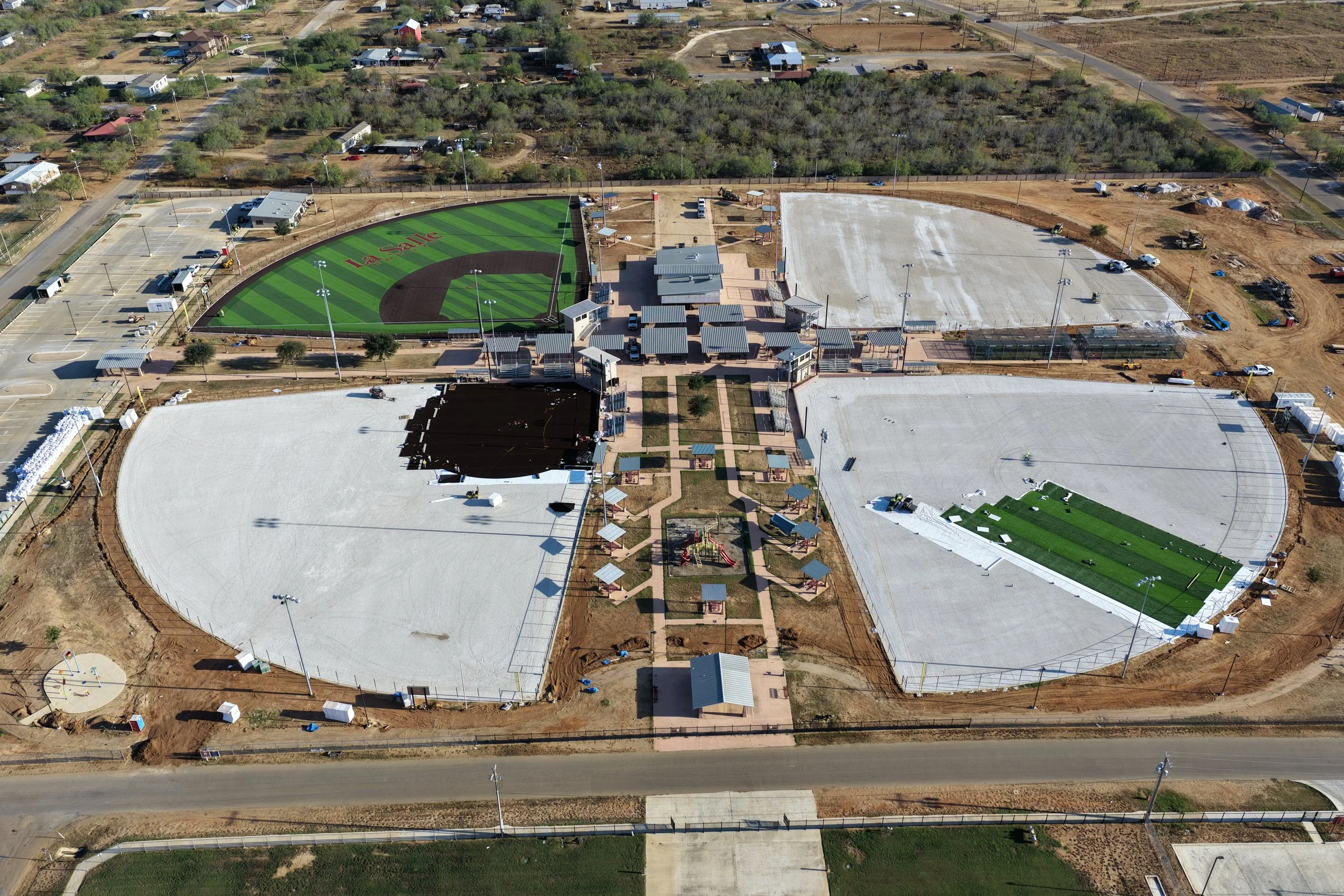 Aerial view of a sports complex with multiple fields, including a baseball field with artificial turf and the words "La Salle" on it, a skate park with skate ramps, a parking lot, and surrounding areas under construction or development.