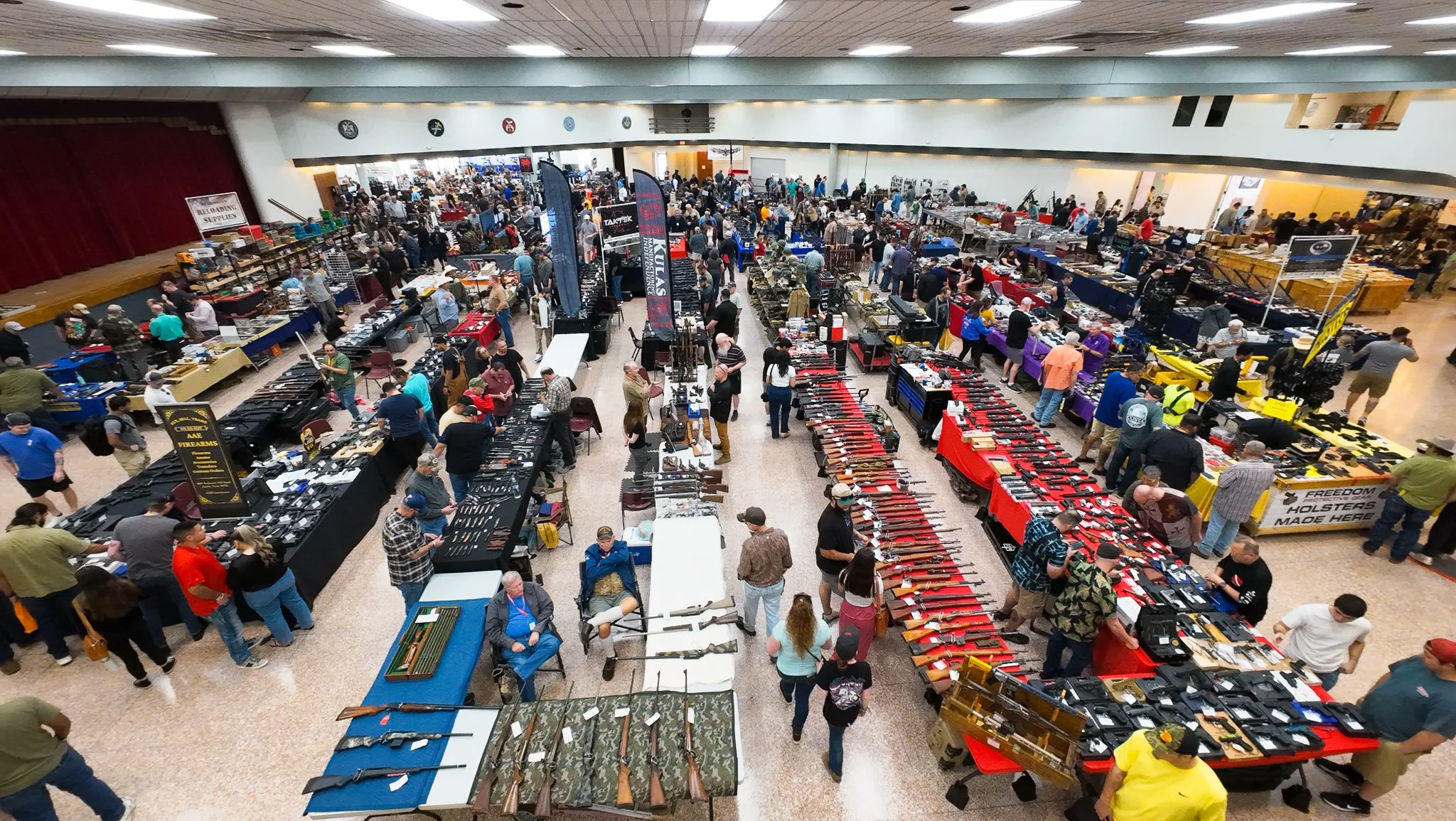 Large indoor gun show with numerous tables displaying firearms, rifles, and weapons; many people browsing and purchasing items; banners for firearm brands and accessories; crowded event.