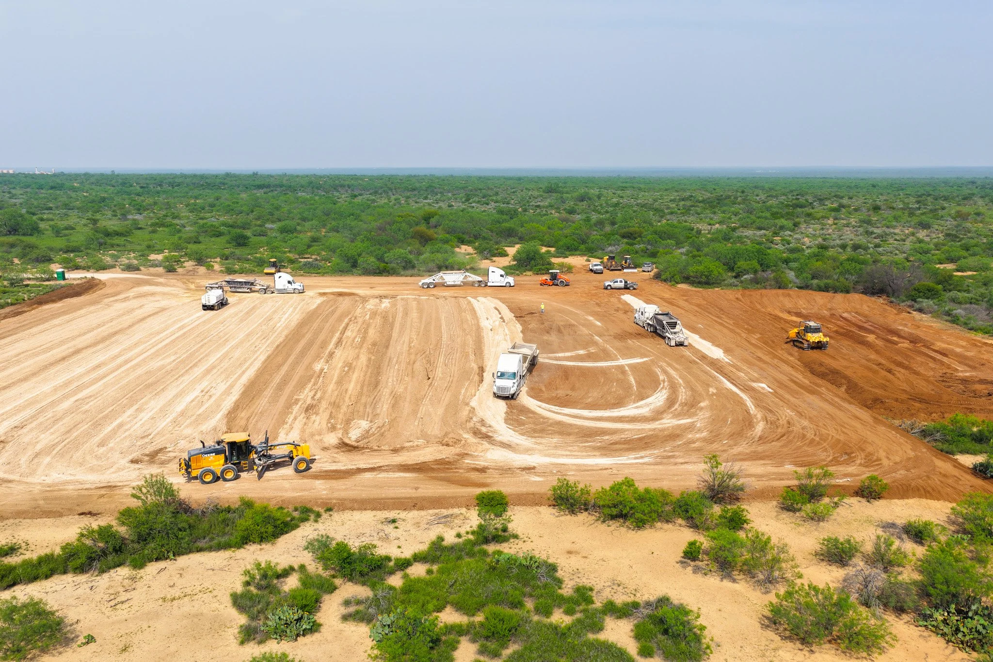 Construction site with various heavy machinery including bulldozers, dump trucks, and pickup trucks working on leveling and preparing the land in a vast green landscape under a cloudy sky.